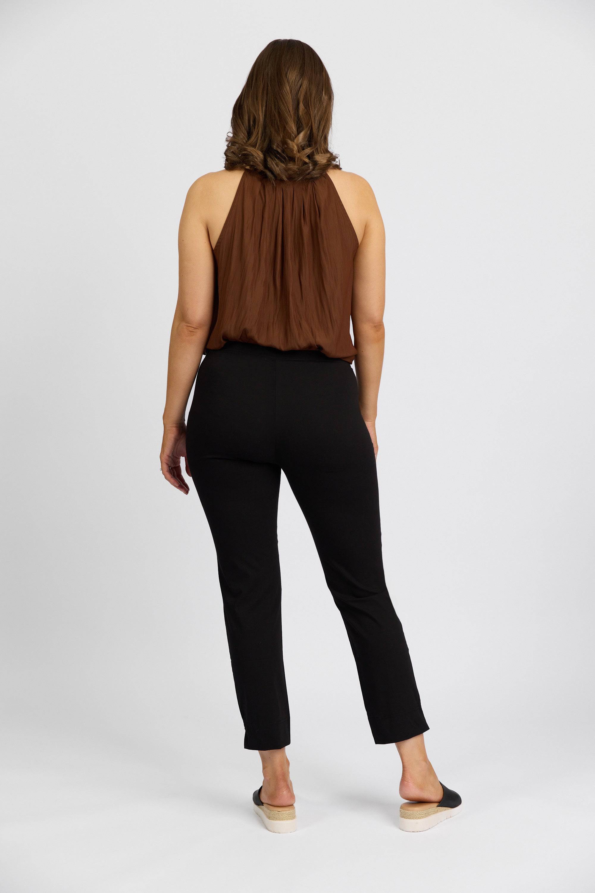A woman with wavy brown hair, wearing a sleeveless brown top and Vassalli Paris Capri Pant - Black, stands facing away from the camera against a plain white background.