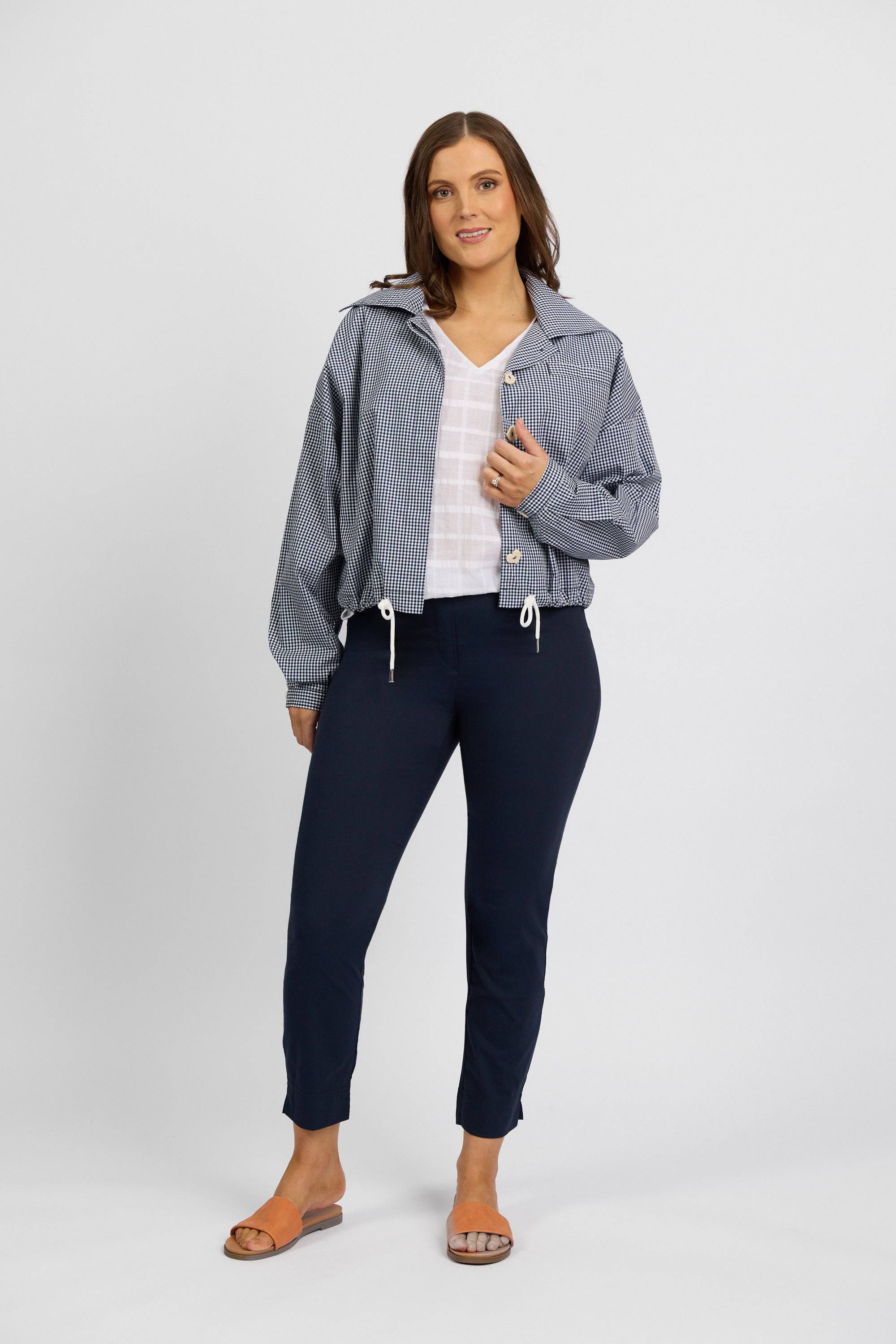 A woman with shoulder-length brown hair stands against a plain background, wearing Vassalli Paris Capri Pant - Inka and a white top. She has tan sandals and a relaxed, confident expression.