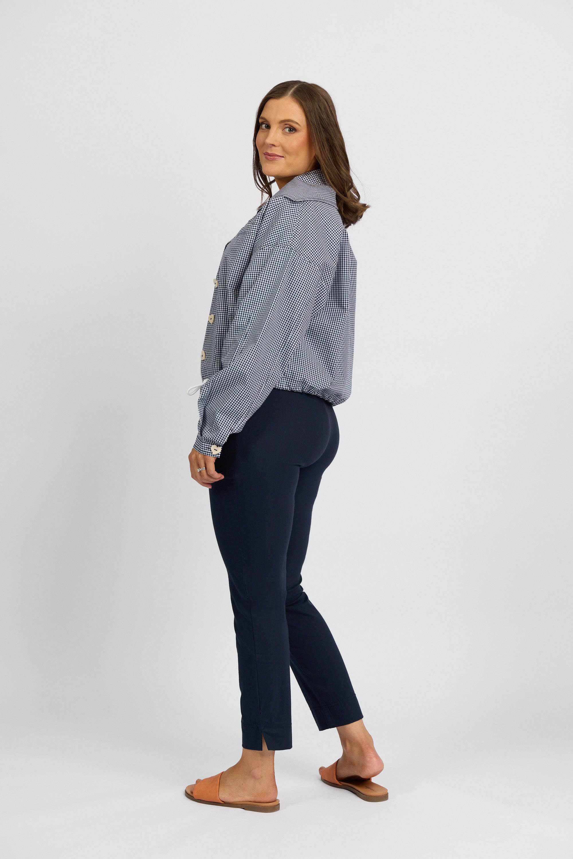 A woman stands sideways, looking at the camera. She wears a blue and white striped blouse, Vassalli Paris Capri Pant - Inka in stretch fabric, and brown sandals against a plain white background.