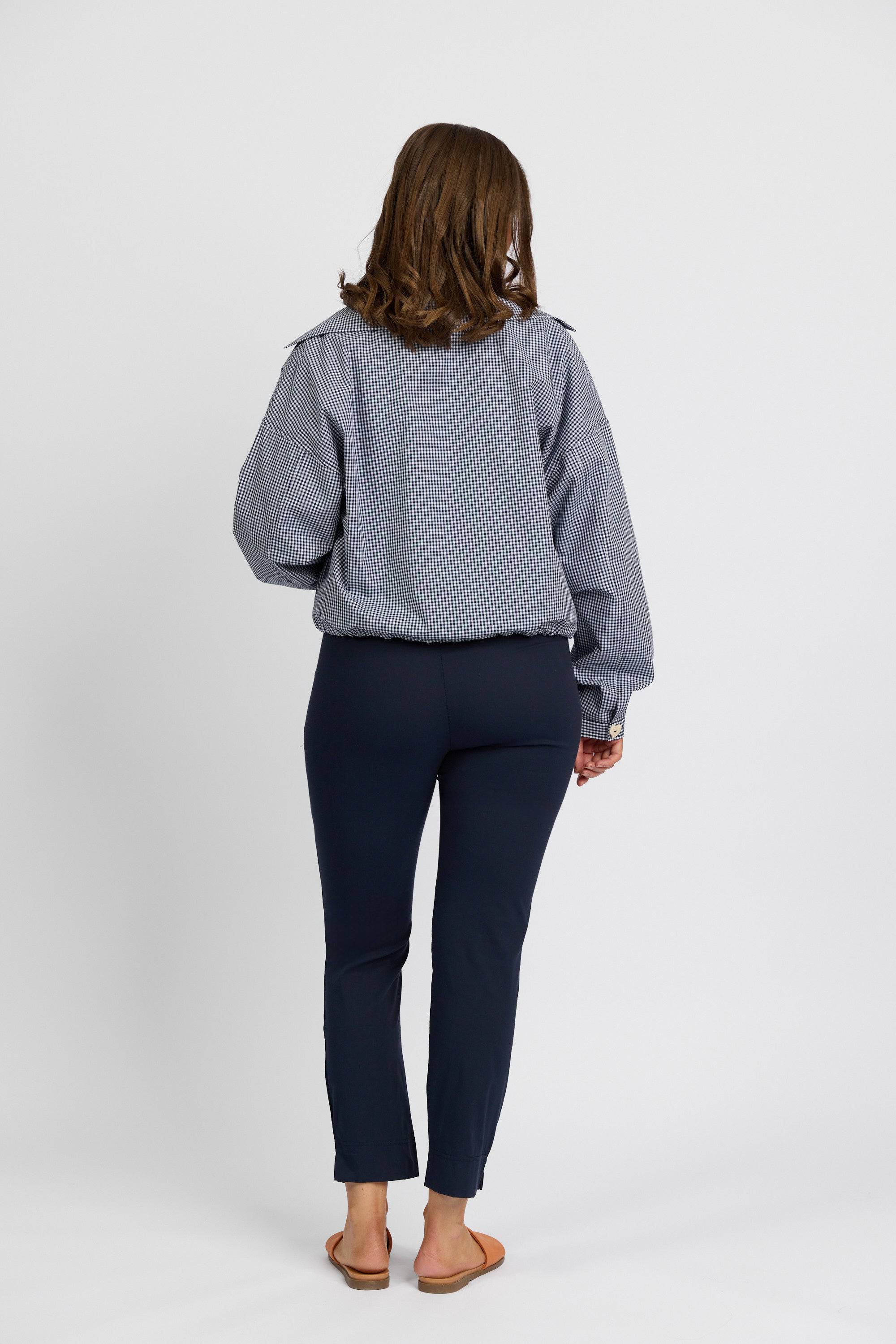 A person with shoulder-length brown hair, seen from behind, wears a blue and white striped long-sleeve shirt, Vassalli Paris Capri Pant - Inka in stretch fabric, and tan sandals, standing against a plain white background.