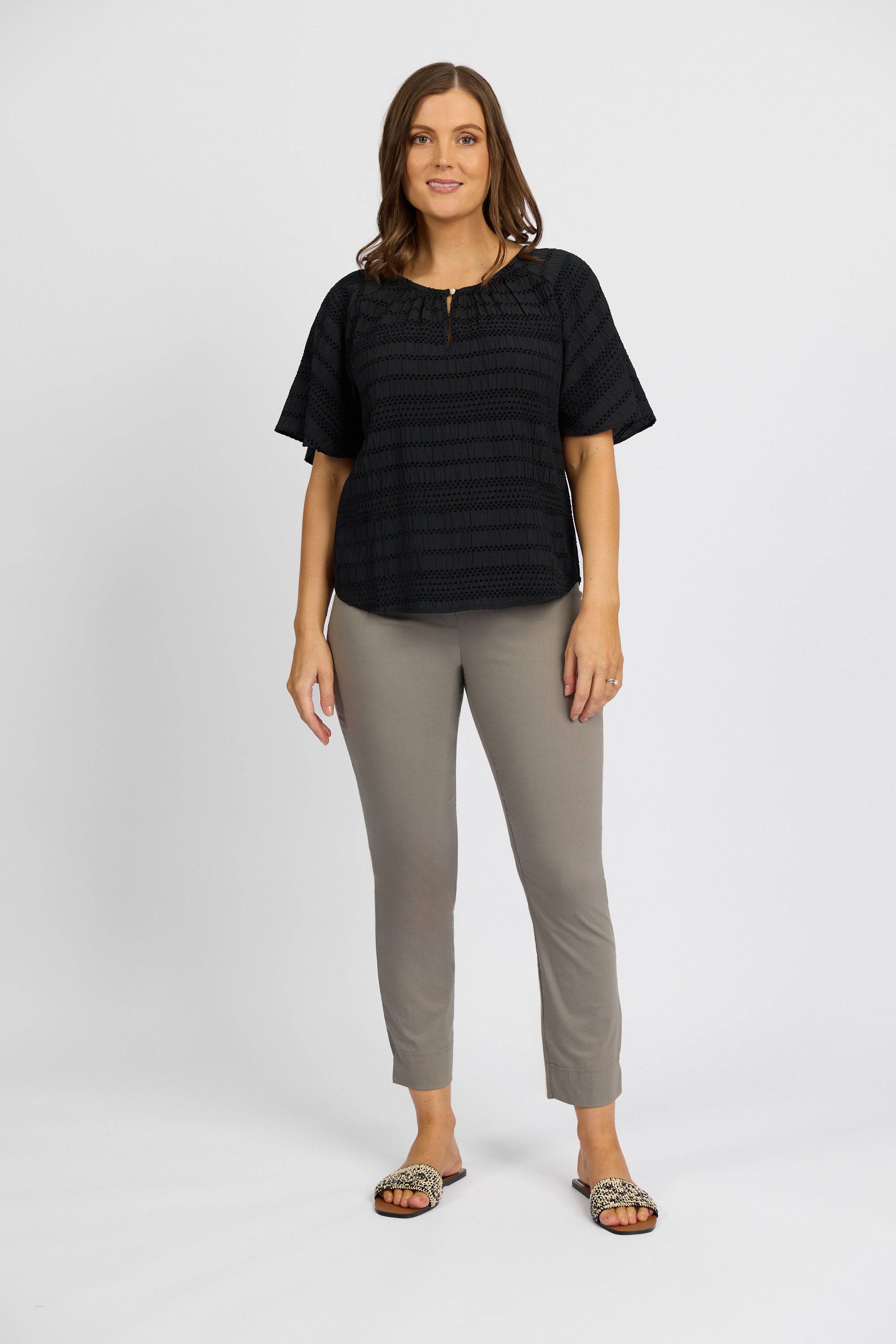 A woman faces forward, smiling, wearing Vassalli Paris Capri Pant - Latte in stretch fabric, a short-sleeve black top, and patterned slip-on sandals. The background is plain white.