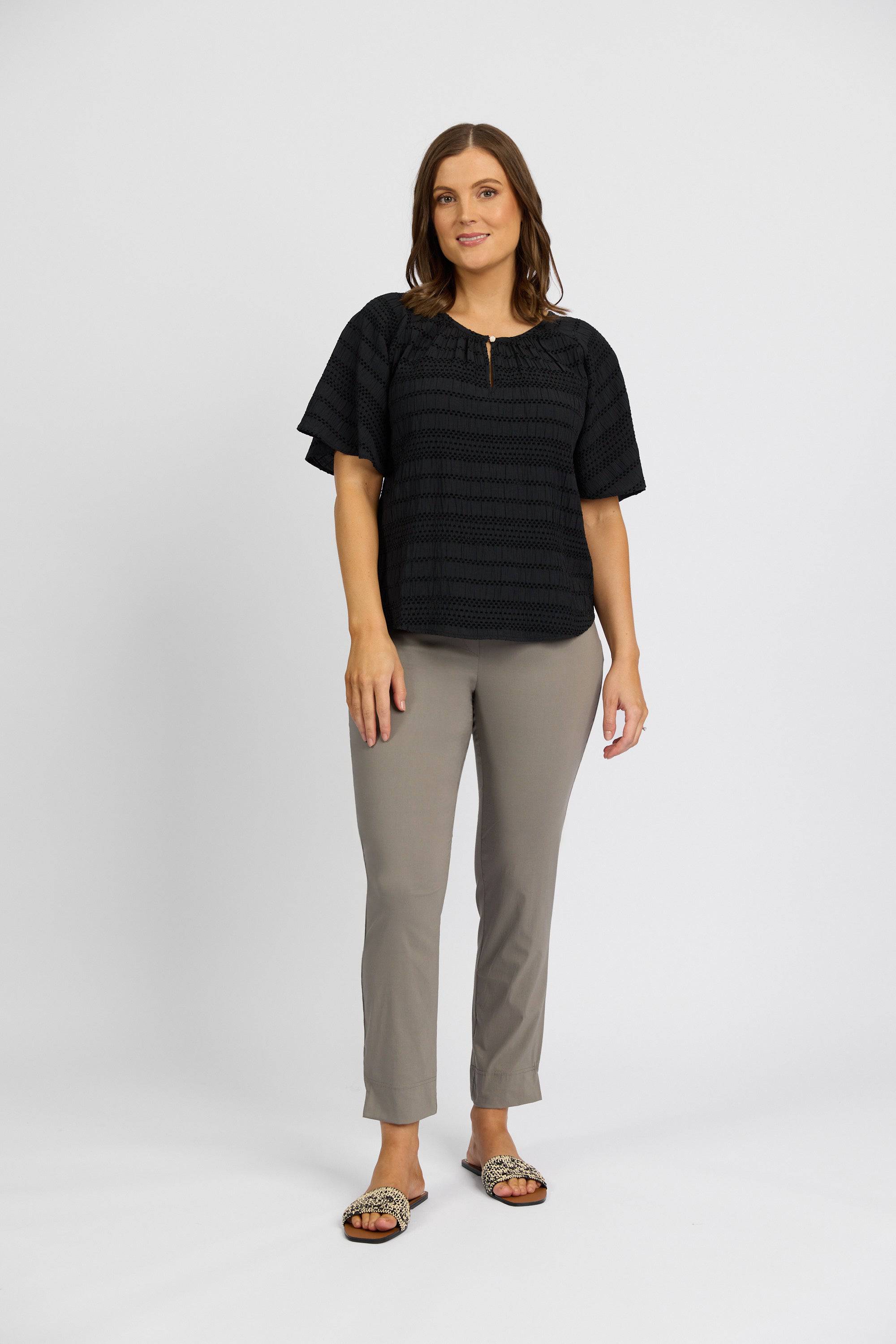 A woman with shoulder-length brown hair and a relaxed smile wears a loose black short-sleeve top, Vassalli Paris Capri Pant - Latte, and open-toe sandals against a plain white background.