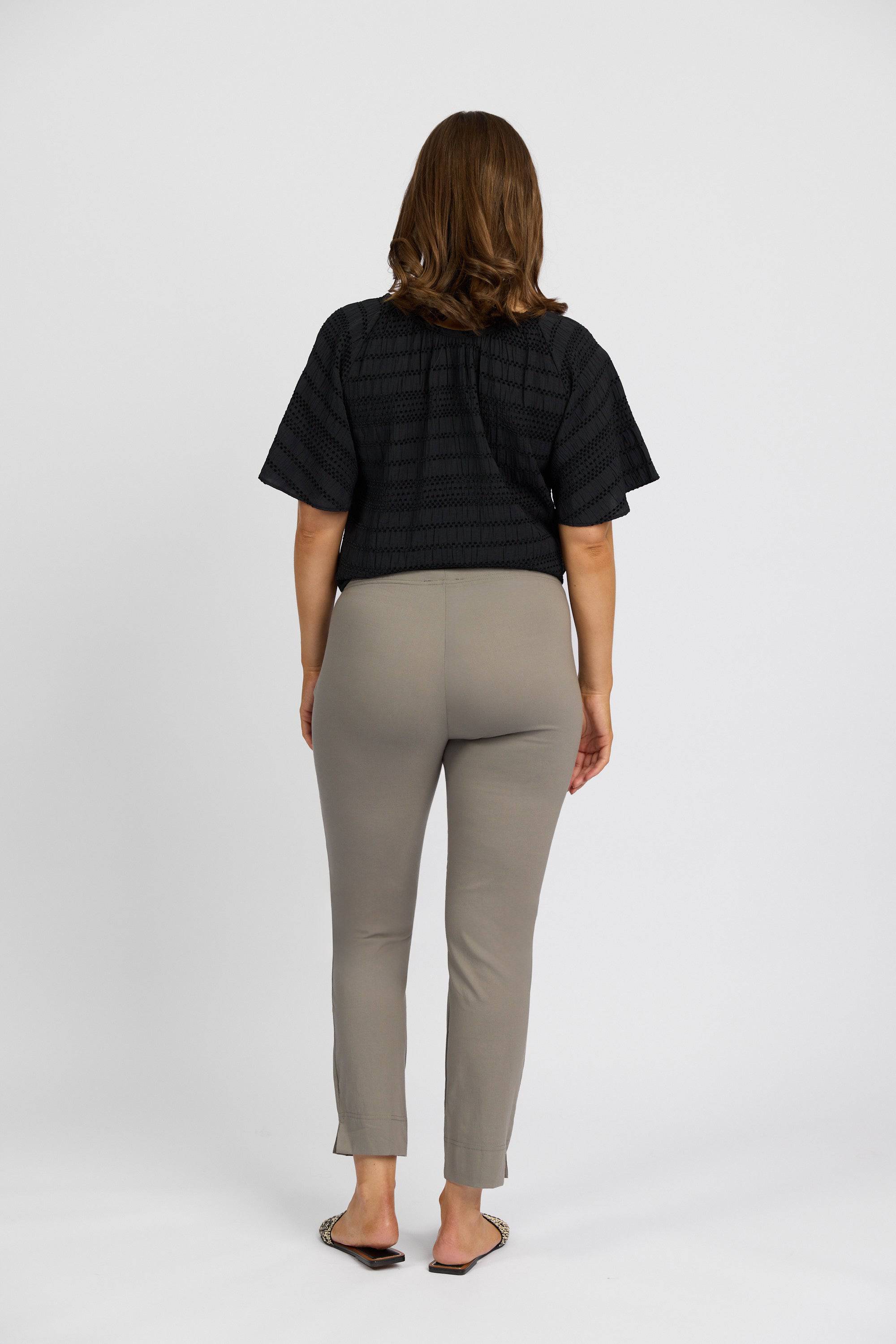 A woman with shoulder-length brown hair, wearing a black short-sleeved top and Vassalli's Paris Capri Pant - Latte made from stretch fabric with a mid rise, stands in flat shoes facing away on a plain white background.