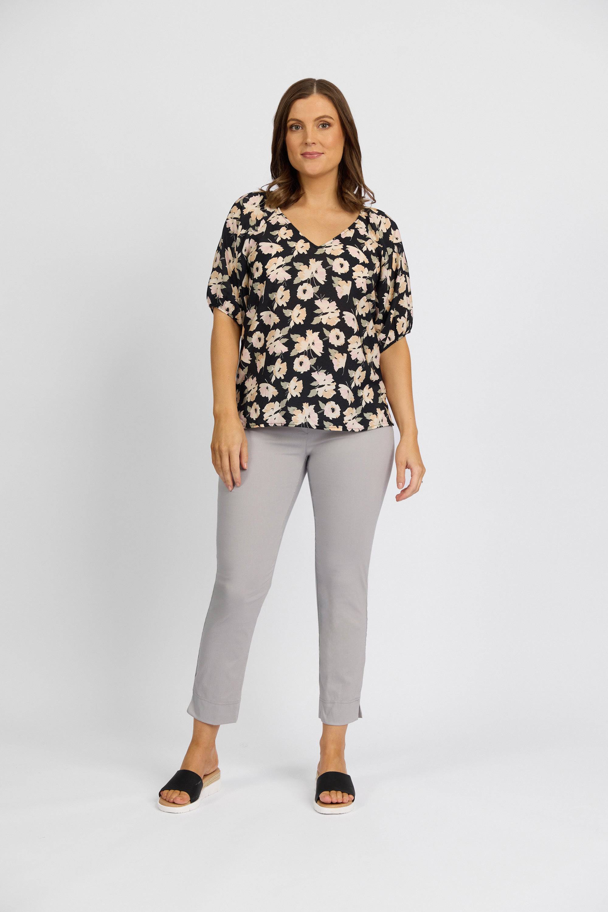 A woman with shoulder-length brown hair and a neutral expression stands against a plain white background, wearing Vassalli’s Paris Capri Pant - Silver in a mid-rise 7/8 fit, paired with a black floral blouse and black slide sandals.