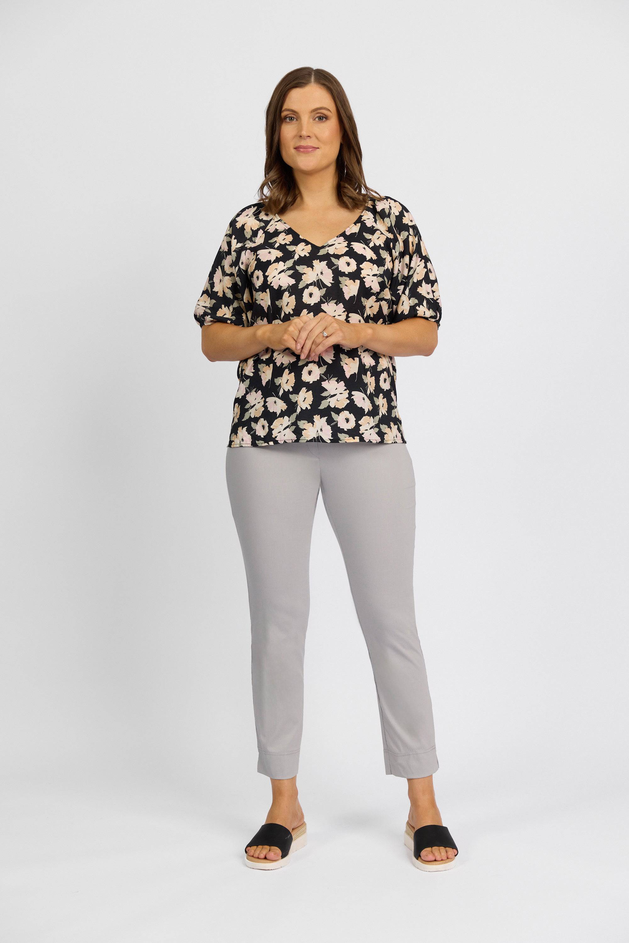 A woman faces forward, wearing a black floral top and Vassalli Paris Capri Pant - Silver with a mid rise 7/8 fit. Her shoulder-length brown hair frames her slight smile as she stands against a plain white background.