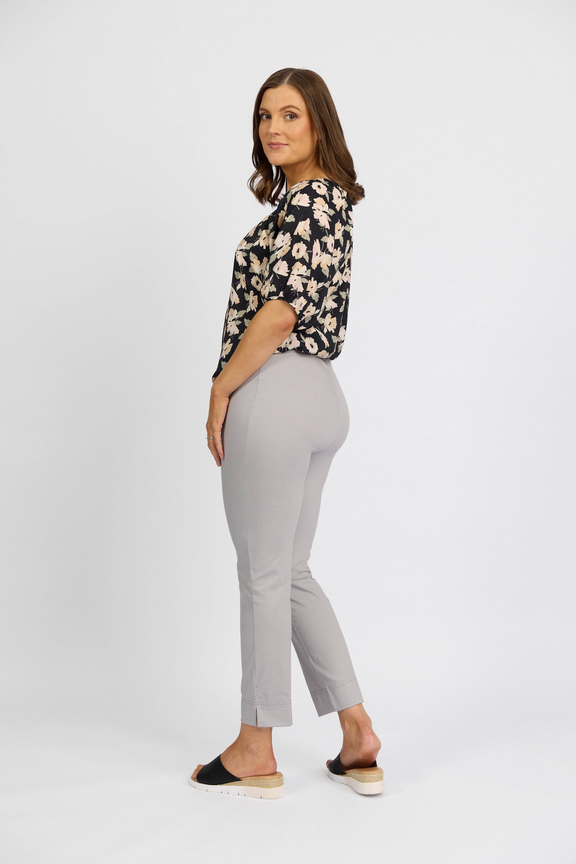 A woman stands sideways, looking at the camera, wearing Vassalli Paris Capri Pant - Silver, a black floral blouse, and black slip-on shoes with a slight platform heel against a plain white background.