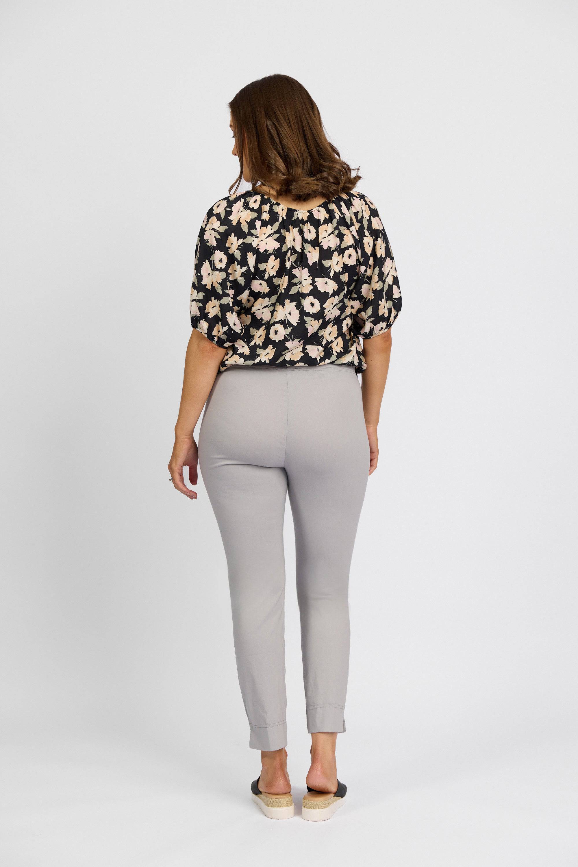 A woman faces away from the camera, wearing a black floral blouse and Vassalli Paris Capri Pant - Silver, crafted from stretch fabric, paired with black slip-on shoes against a plain white background.