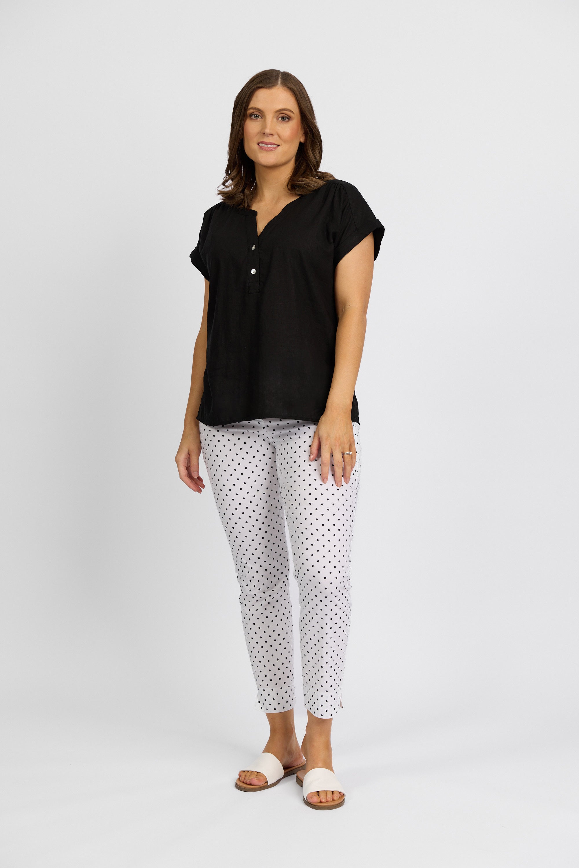 A woman with shoulder-length hair and a relaxed expression wears Vassalli's Paris Printed Capri Pant - Spot, a mid-rise 7/8 black polka dot style, paired with a black short-sleeve blouse and white slide sandals against a plain white background.