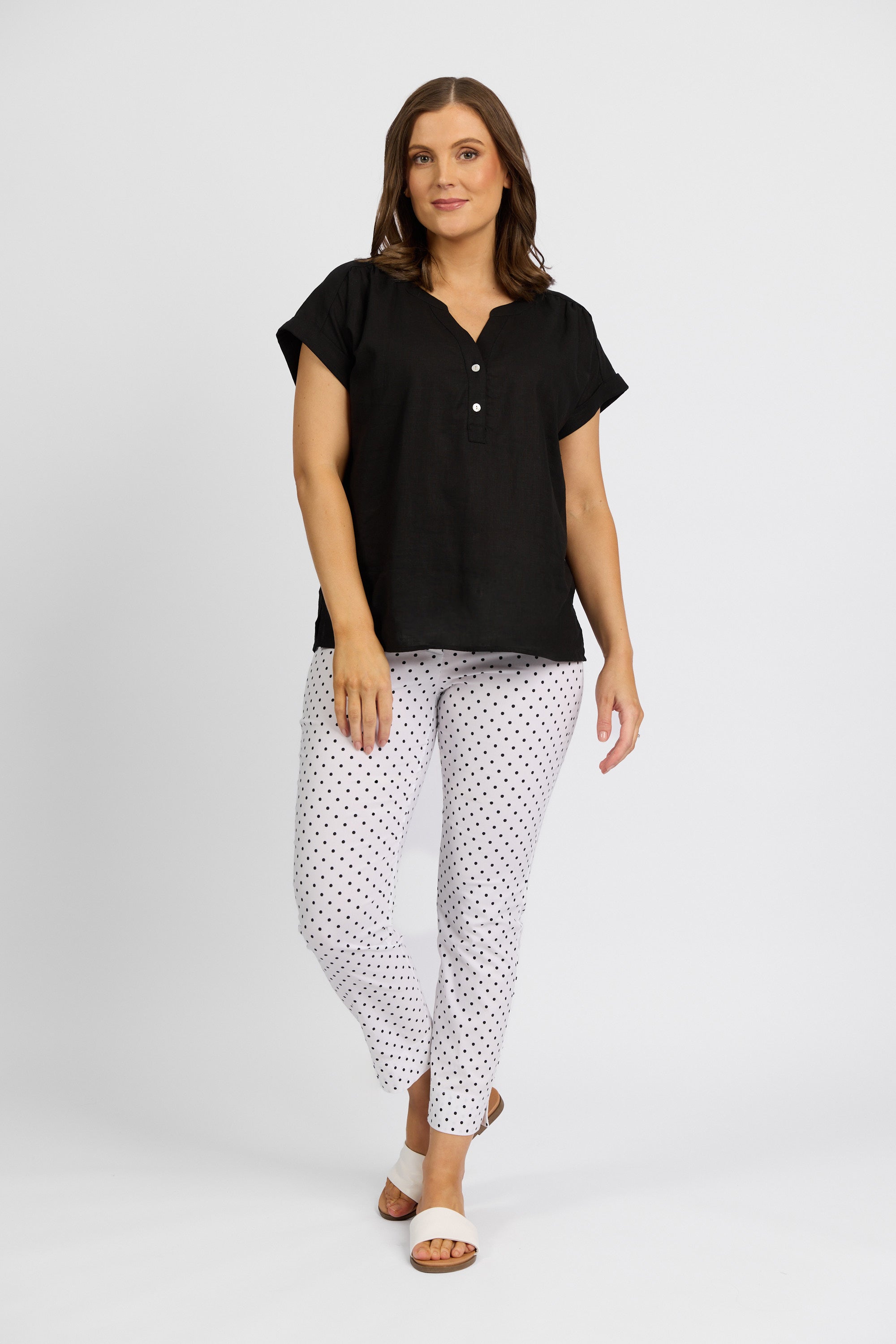 A woman with shoulder-length brown hair stands against a white background, wearing a loose black top, Vassalli Paris Printed Capri Pant - Spot in stretch white fabric with black dots, and white sandals. She looks relaxed.