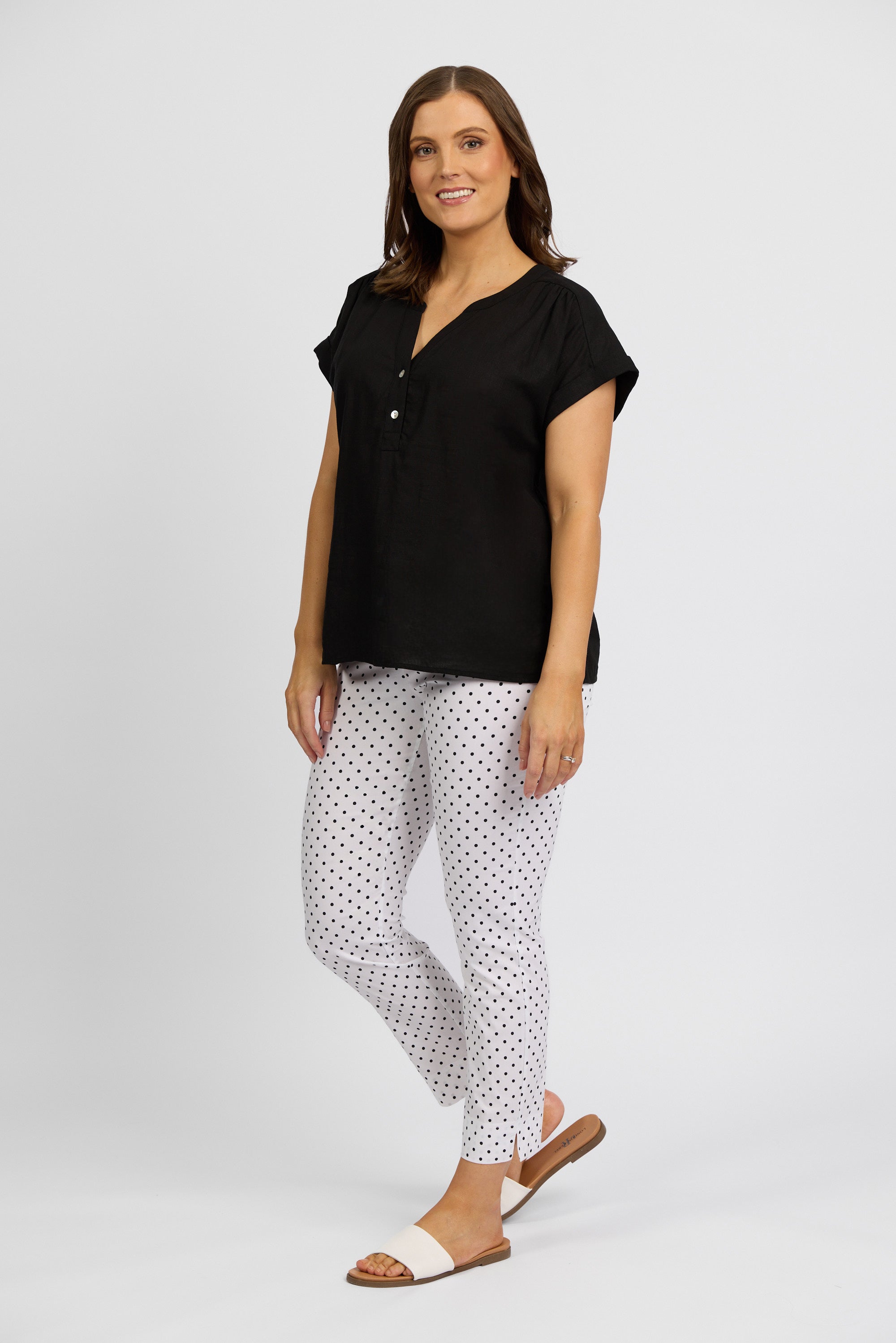 A woman stands against a plain background, smiling with one leg slightly bent. She wears a black short-sleeve blouse and Vassalli Paris Printed Capri Pant - Spot, paired with brown-and-white sandals.
