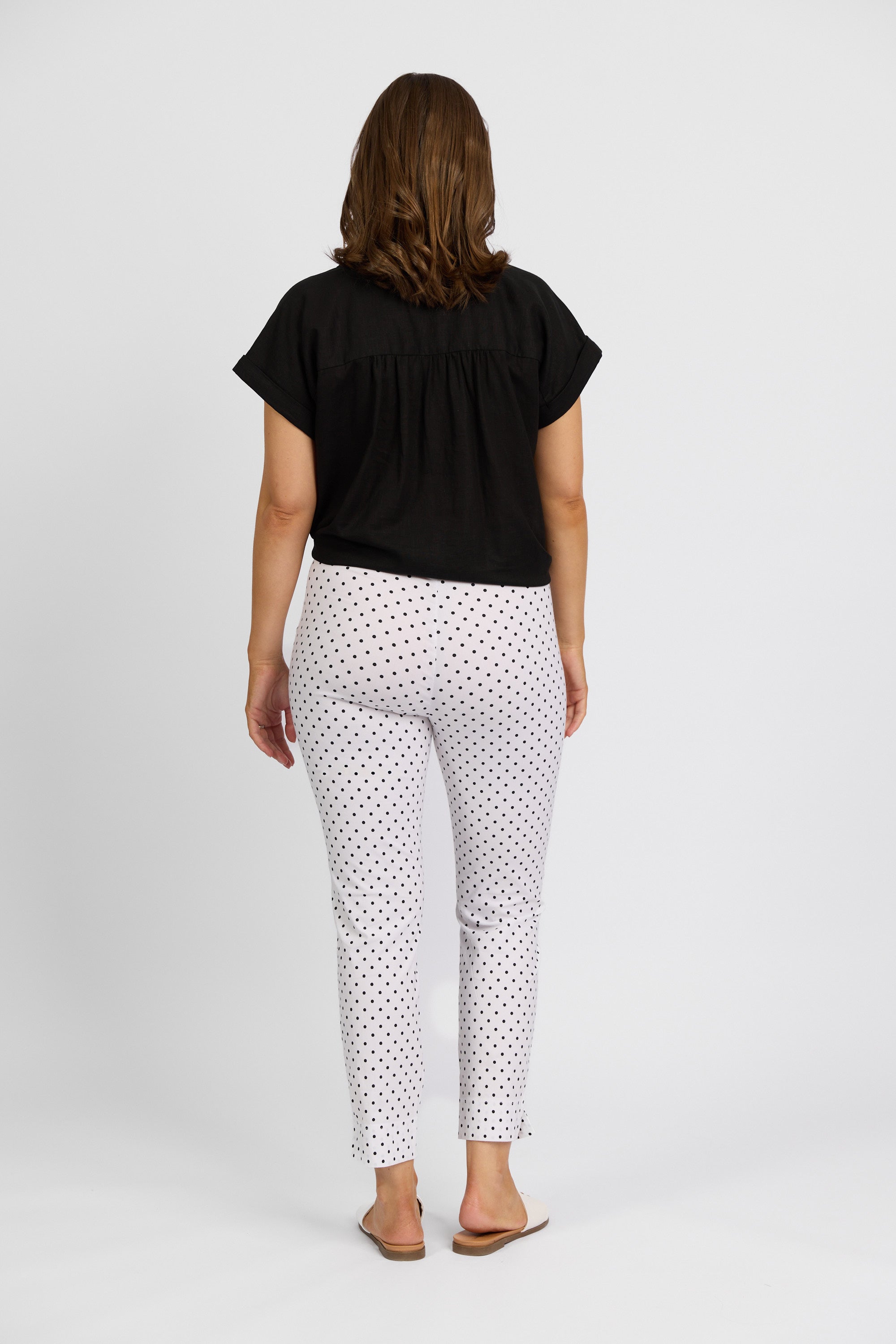 A woman faces away from the camera, wearing a black short-sleeve top and Vassalli Paris Printed Capri Pant - Spot in a mid-rise 7/8 fit, paired with beige flats, set against a plain white background.