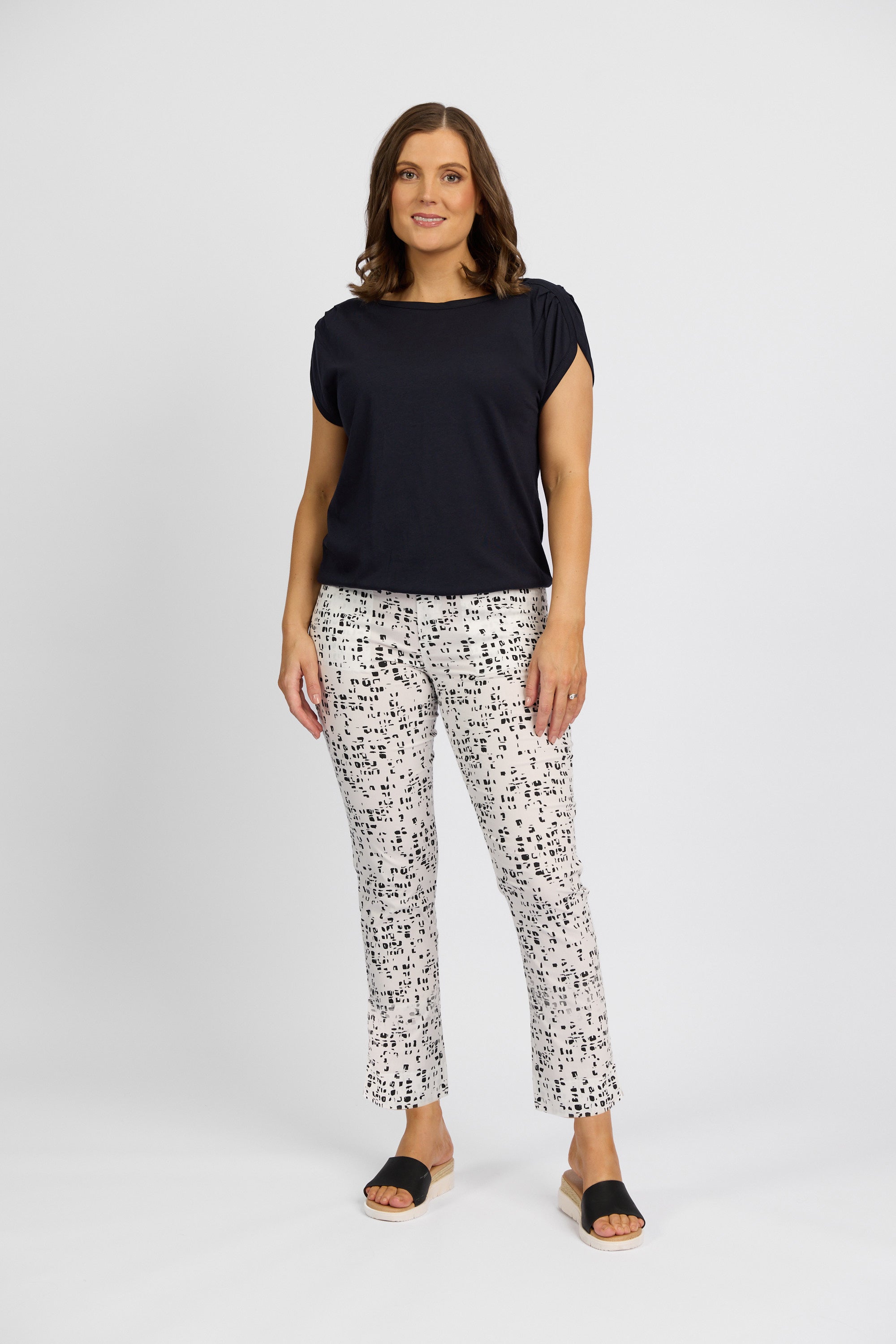 A woman with shoulder-length brown hair smiles against a white background, wearing a black short-sleeve top, Vassalli Paris Printed Capri Pant - Tonic with a black abstract pattern, and black open-toe sandals.