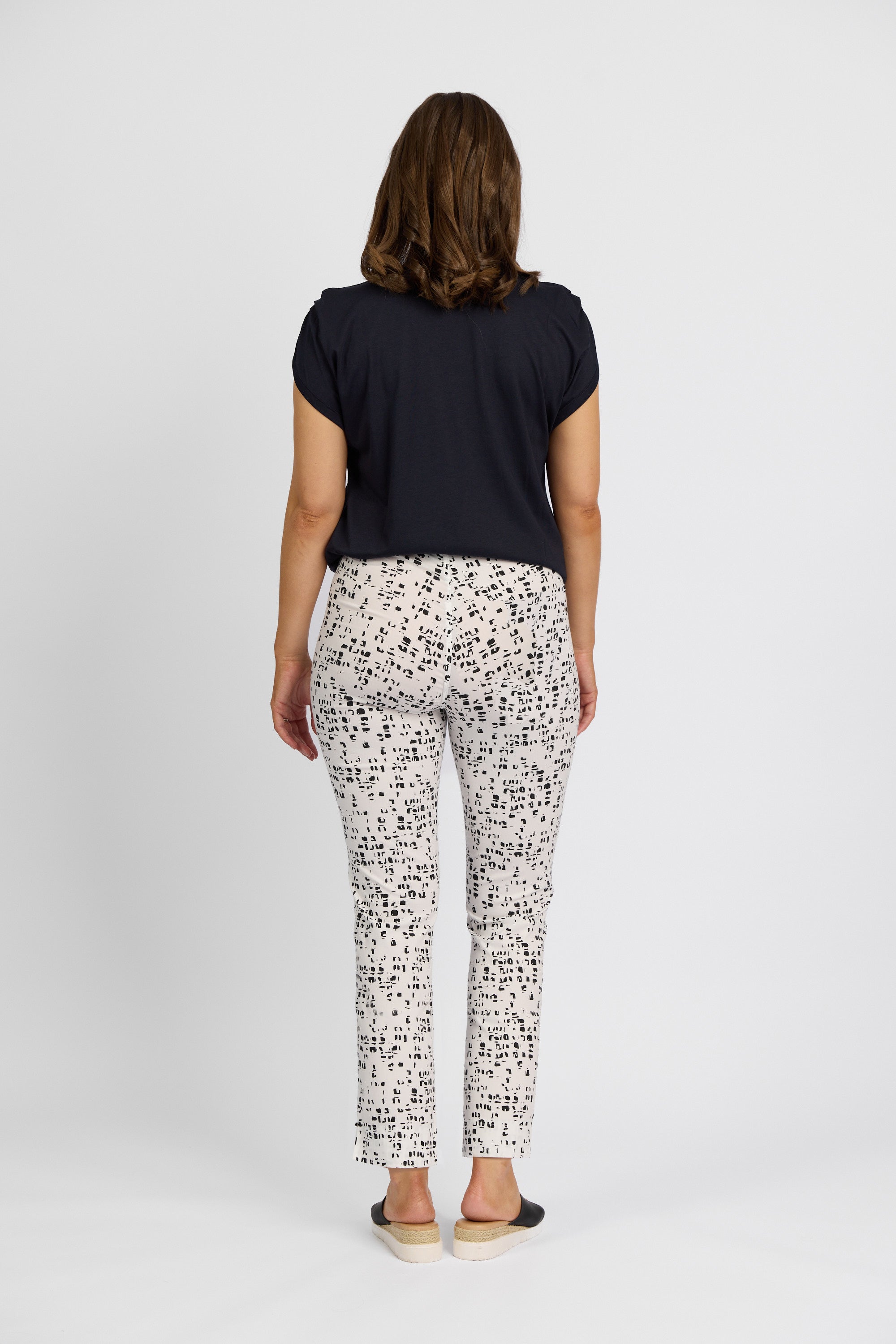 A woman models the Vassalli Paris Printed Capri Pant - Tonic, featuring a black abstract square pattern, paired with a black short-sleeved top and flats, from the Paris Collection, standing against a plain white background.