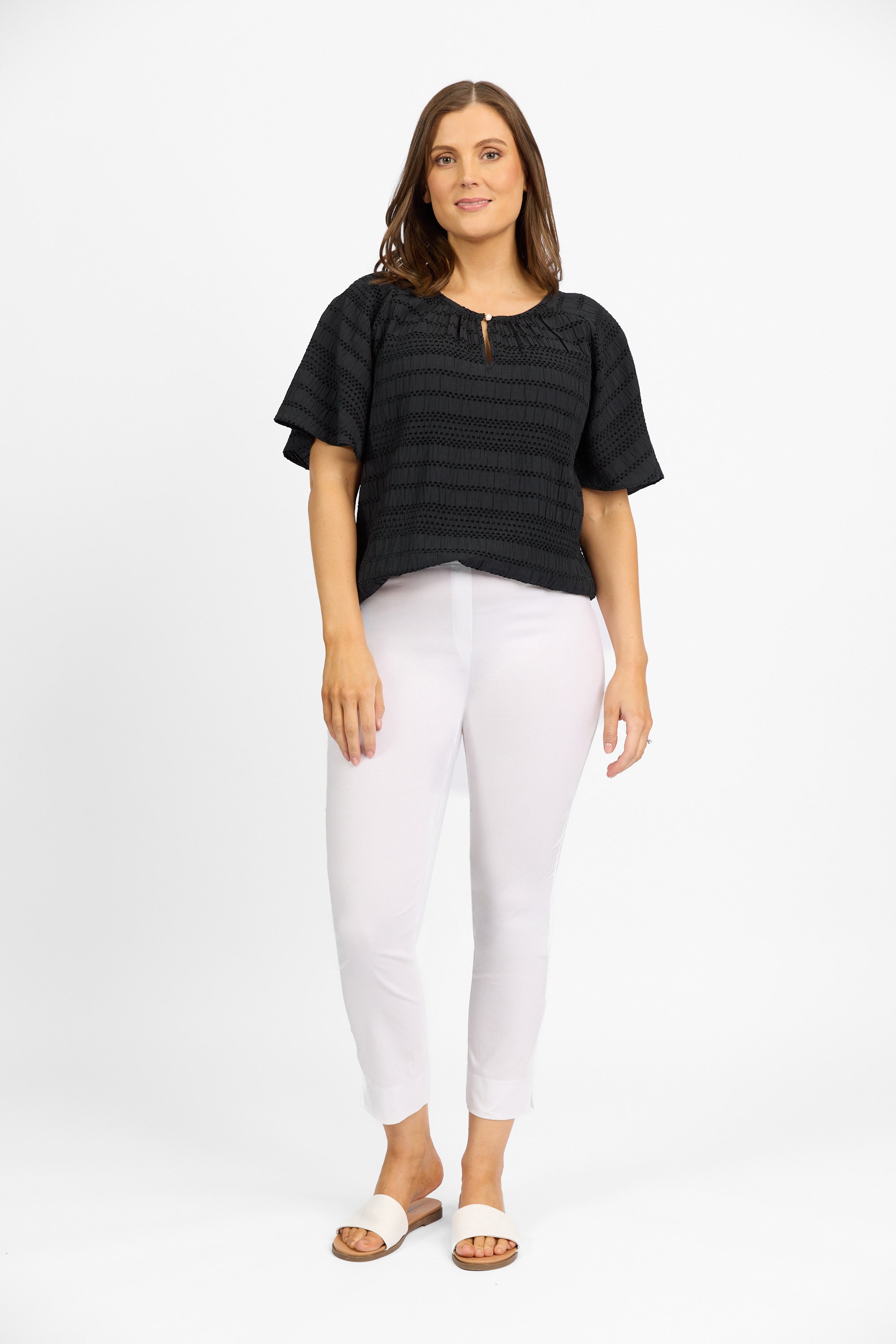 A woman with long brown hair poses confidently against a white background, wearing a Vassalli Paris Capri Pant in white, a black short-sleeve top, and tan slide sandals.