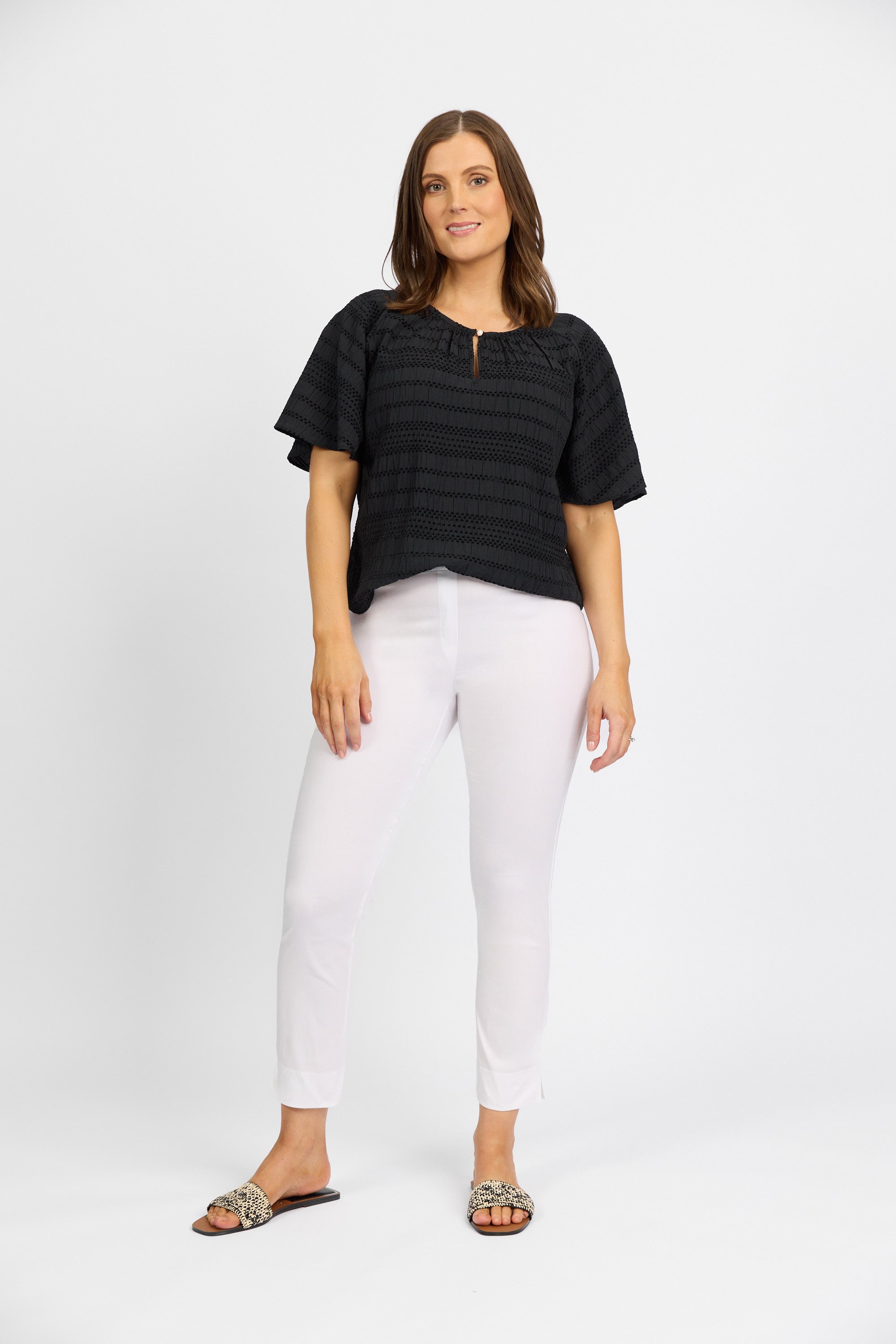 A woman with straight brown hair wears a loose black short-sleeve top, Vassalli Paris Capri Pant - White, and leopard-print slide sandals against a white background, showing a relaxed and confident expression.