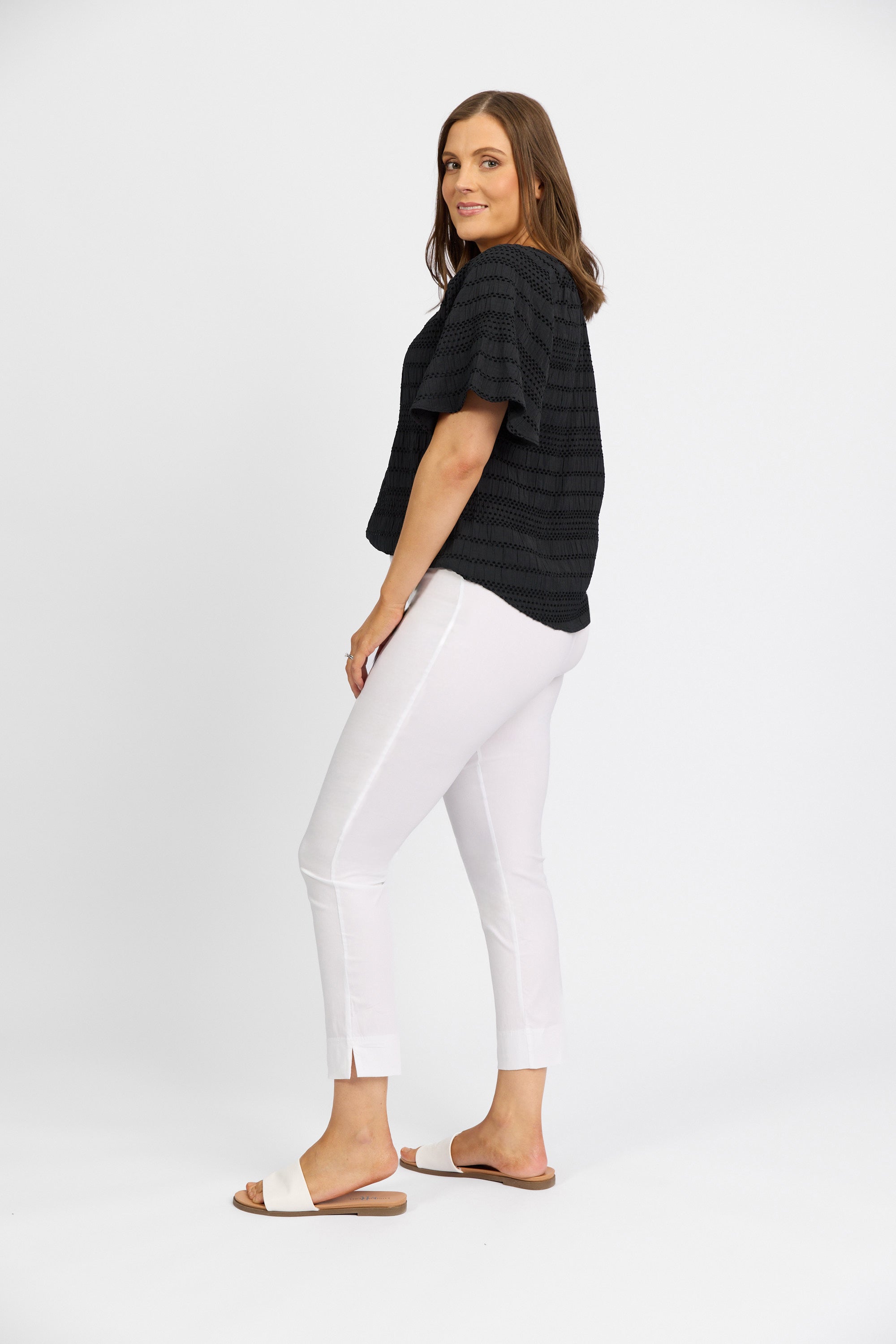 A woman stands sideways against a white background, wearing Vassalli’s Paris Capri Pant in white with a black short-sleeve top and white open-toe sandals. She looks over her shoulder with a slight smile.