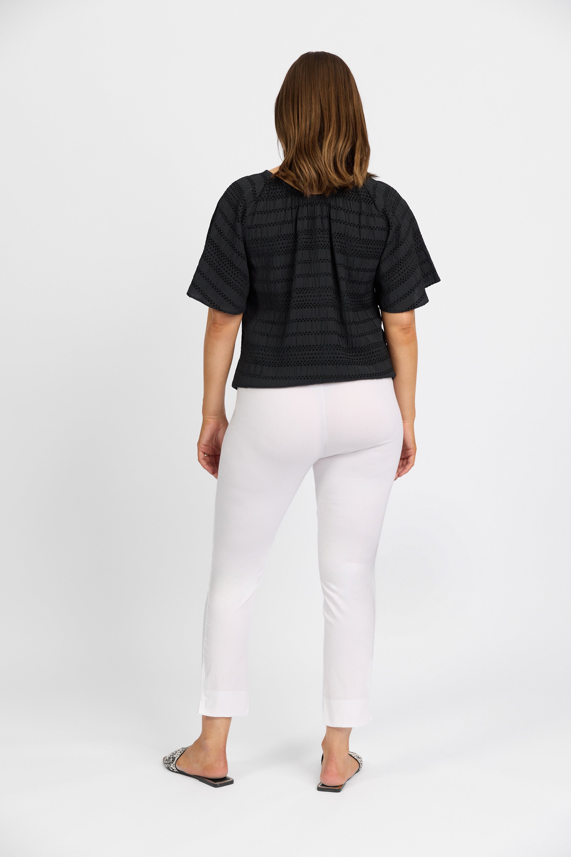 A woman with shoulder-length brown hair faces away from the camera, wearing a black short-sleeve top and Vassalli Paris Capri Pant in White made from stretch fabric, posing against a plain white background.