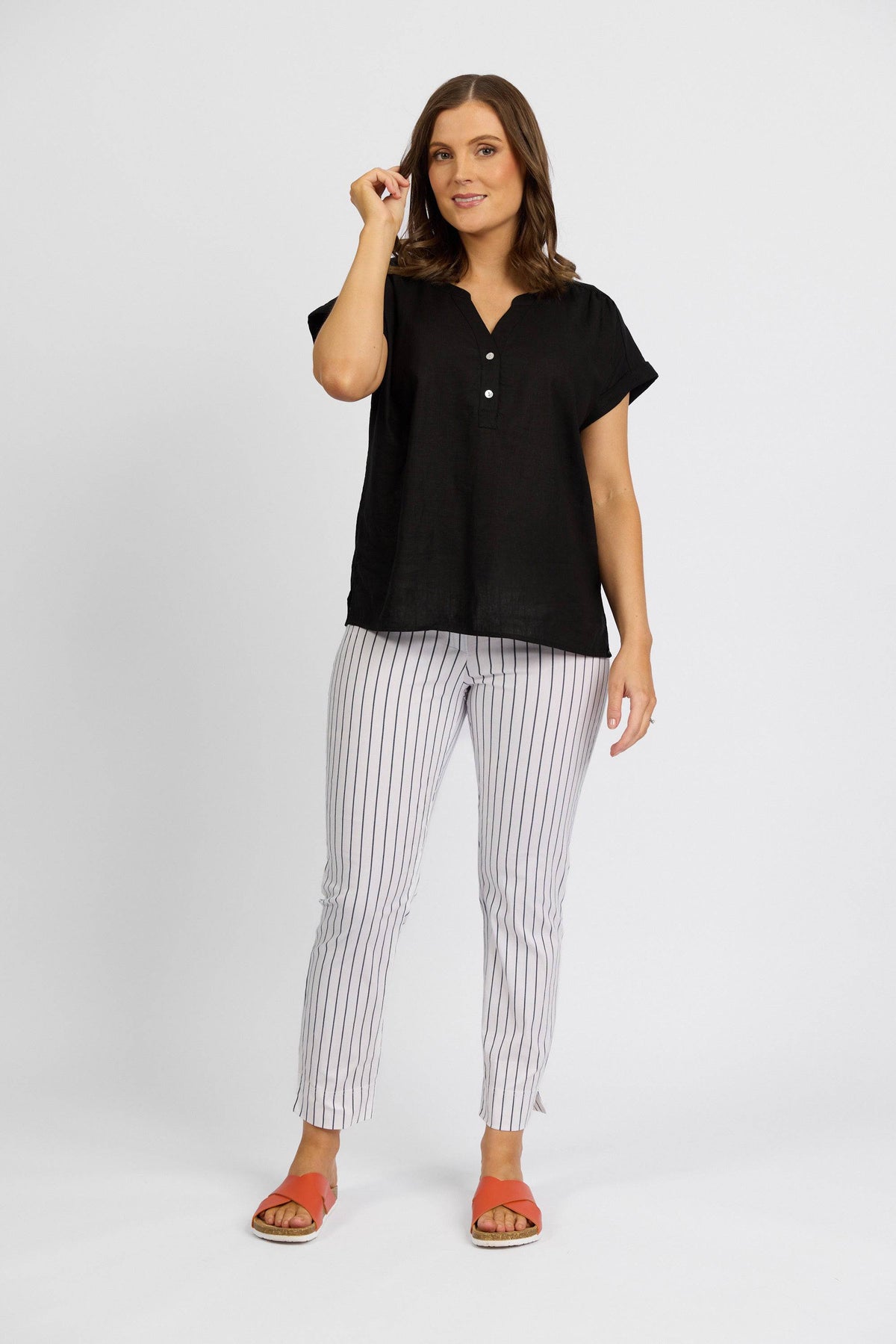 A woman stands against a white background, smiling as she touches her hair. She wears a black short-sleeve blouse, Vassalli Paris Printed Capri Pant in white with stripes, and orange sandals.
