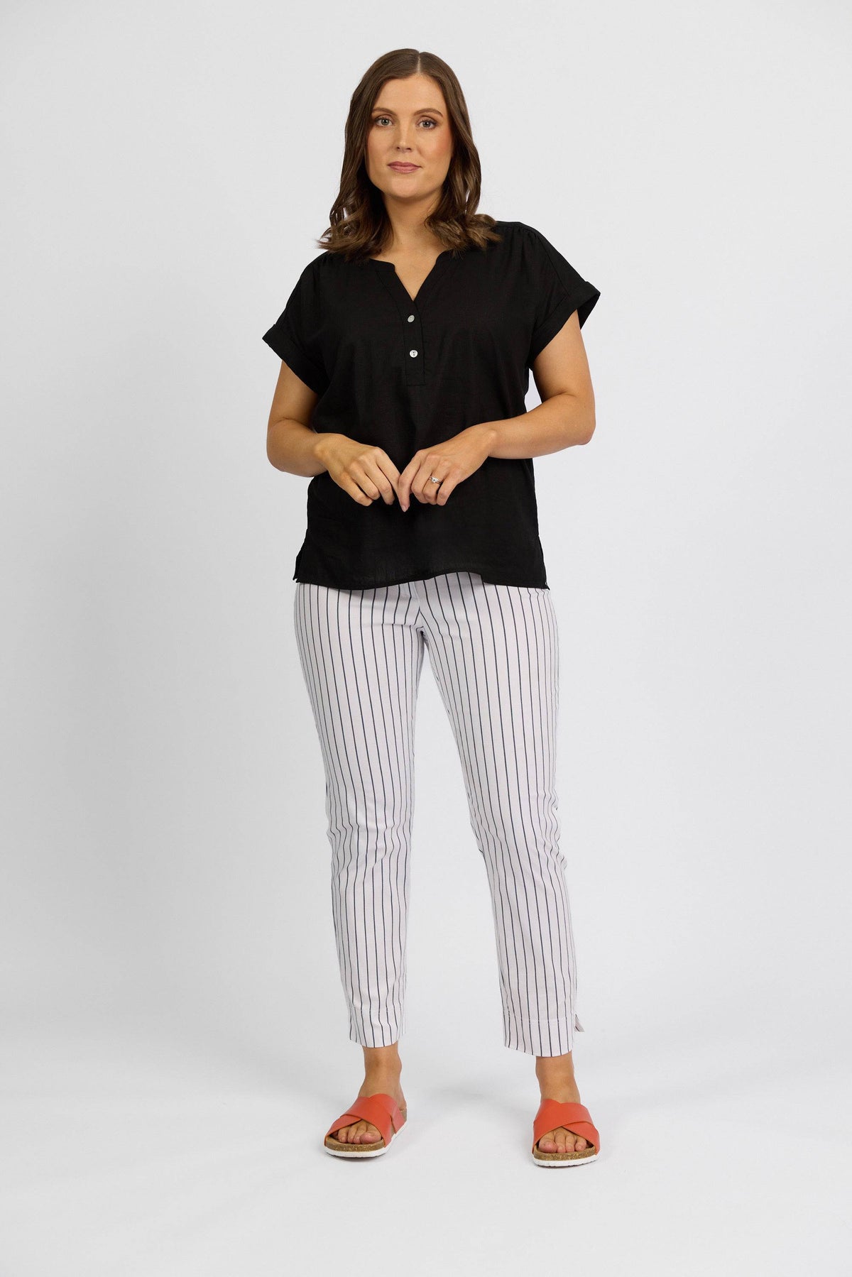 A woman with shoulder-length brown hair stands against a plain background, wearing a black short-sleeve top and Vassalli Paris Printed Capri Pant in white with vertical black stripes. She pairs the look with coral sandals and looks at the camera neutrally.