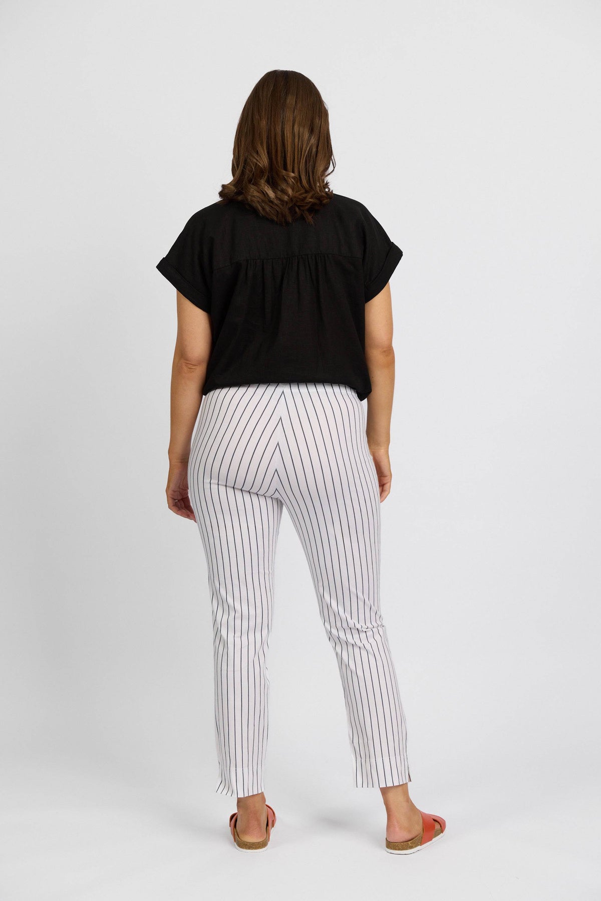 A woman faces away from the camera, wearing a black short-sleeve top and Vassalli Paris Printed Capri Pant - White w/ Stripe in a mid-rise 7/8 fit, paired with orange sandals, against a plain white background.
