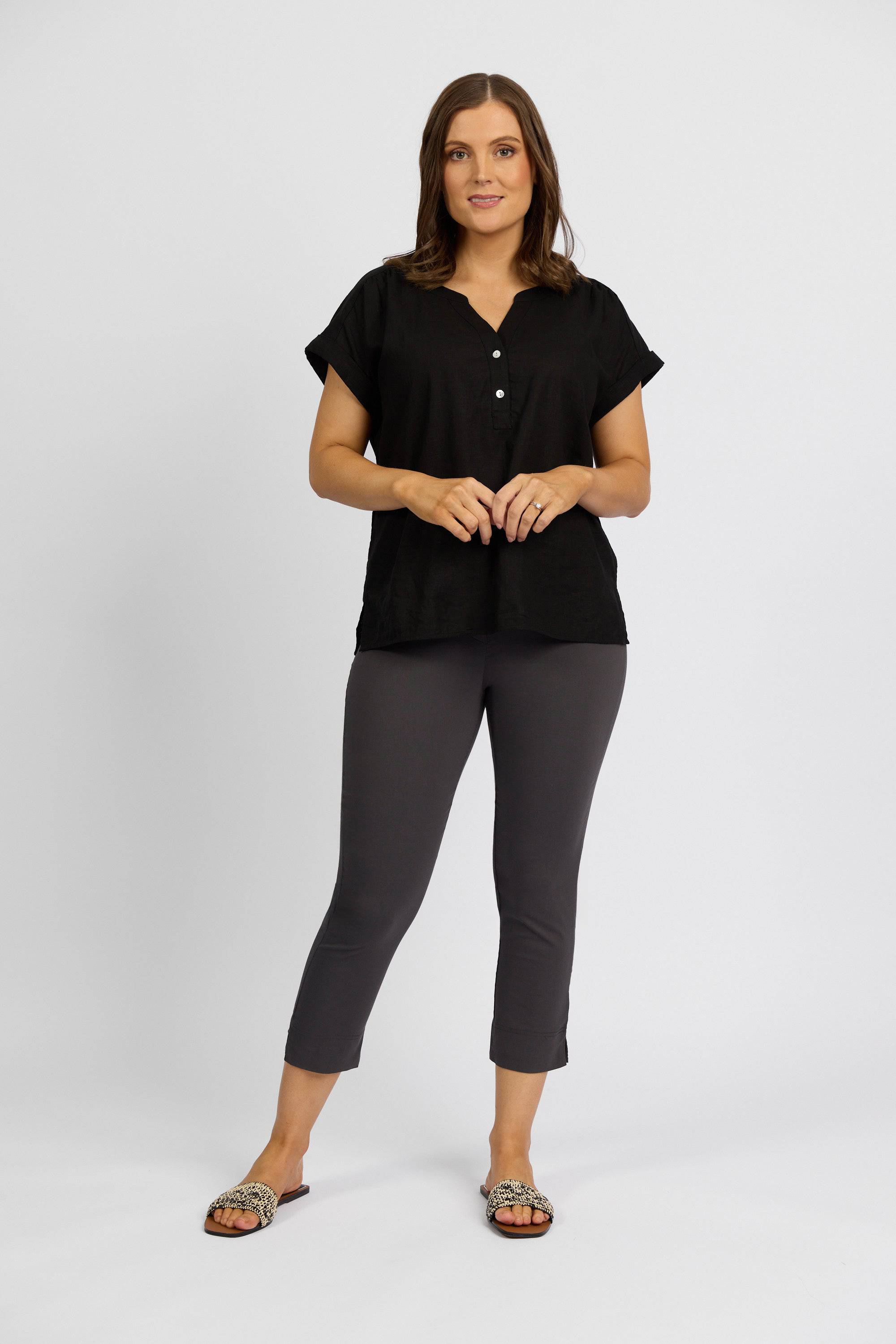 A woman stands against a plain white background wearing a black short-sleeve buttoned top, Vassalli Paris Crop Pant - Beluga, and patterned slide sandals. She faces forward with a relaxed expression and hands together.
