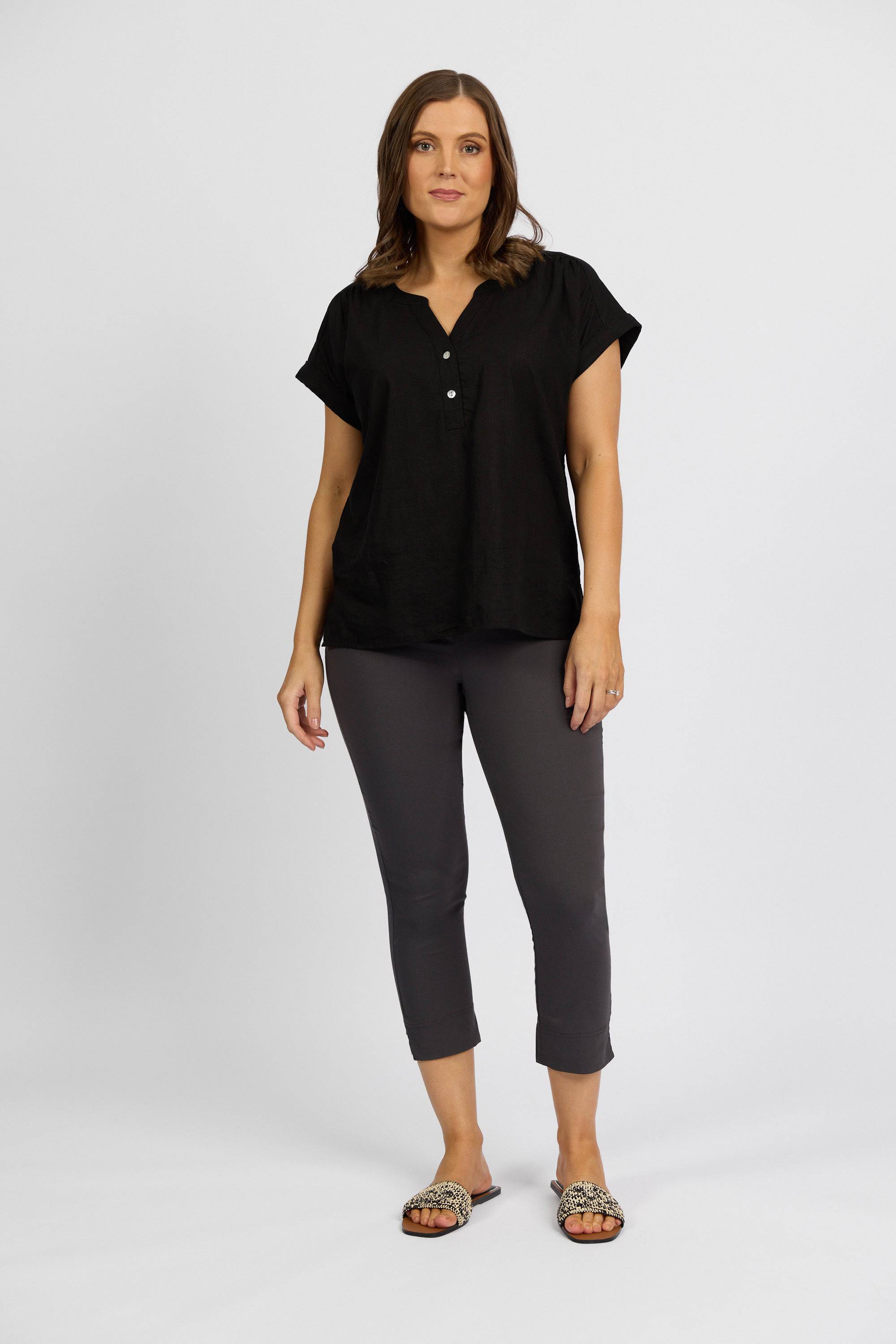 A woman with medium-length hair and a neutral expression stands against a white background, wearing a black short-sleeved blouse, Vassalli Paris Crop Pant - Beluga, and leopard-print slide sandals.