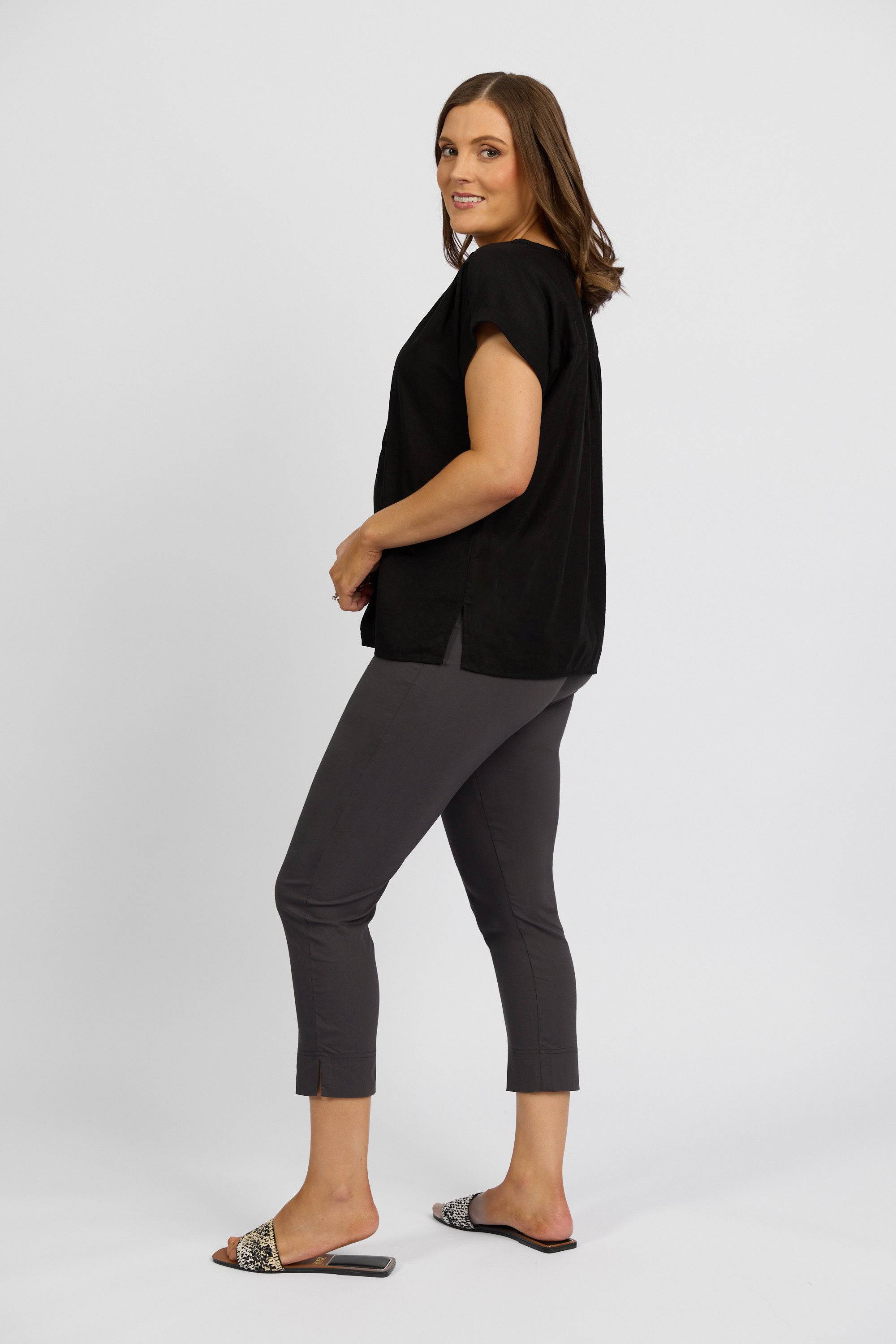 A woman with brown hair wears a loose black short-sleeve top, Vassalli Paris Crop Pant - Beluga, and patterned slide sandals as she stands sideways and smiles at the camera against a plain white background.