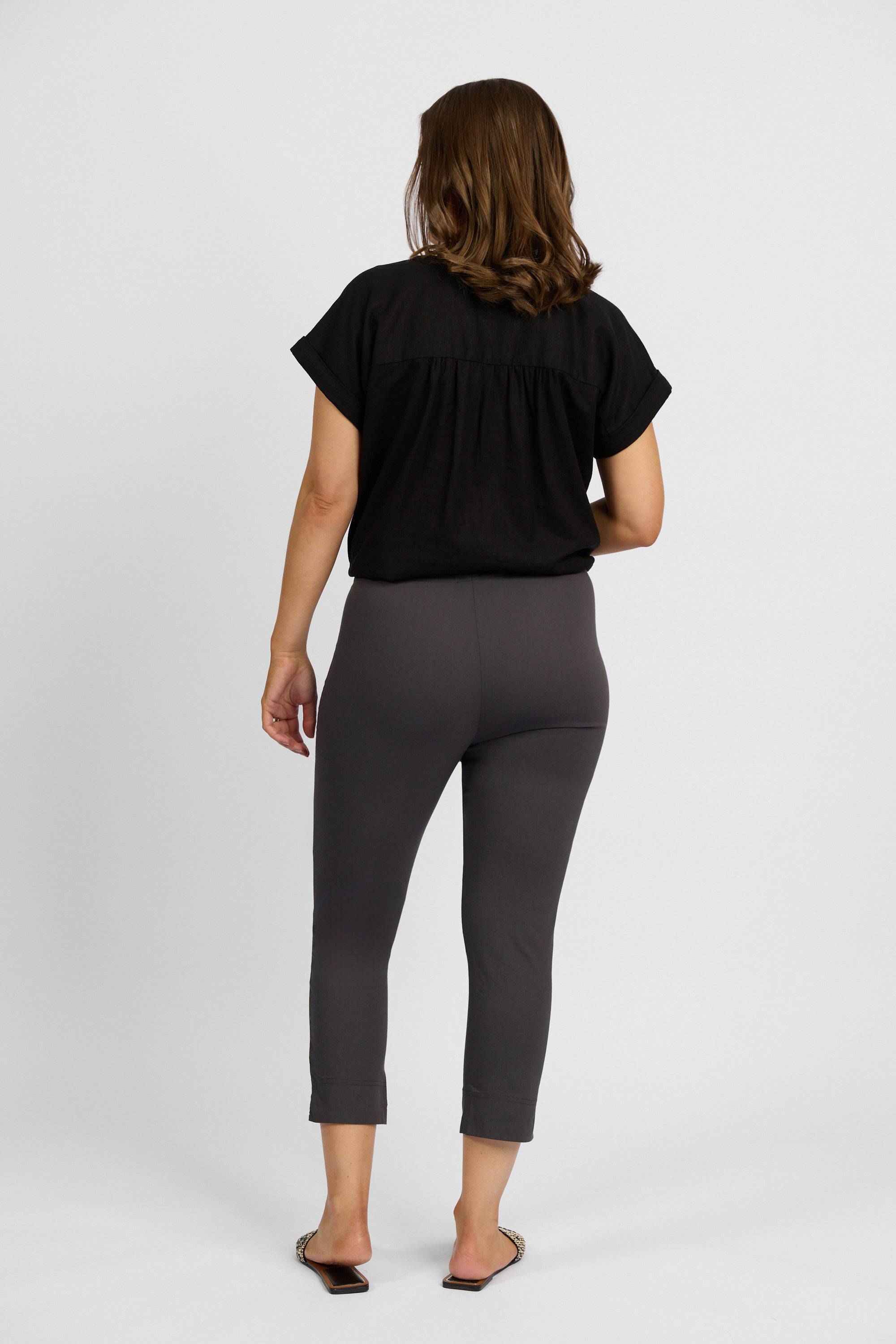 A woman with shoulder-length brown hair faces away from the camera, wearing a black short-sleeve top and Vassalli Paris Crop Pant - Beluga, made in New Zealand from stretch fabric, against a plain white background.