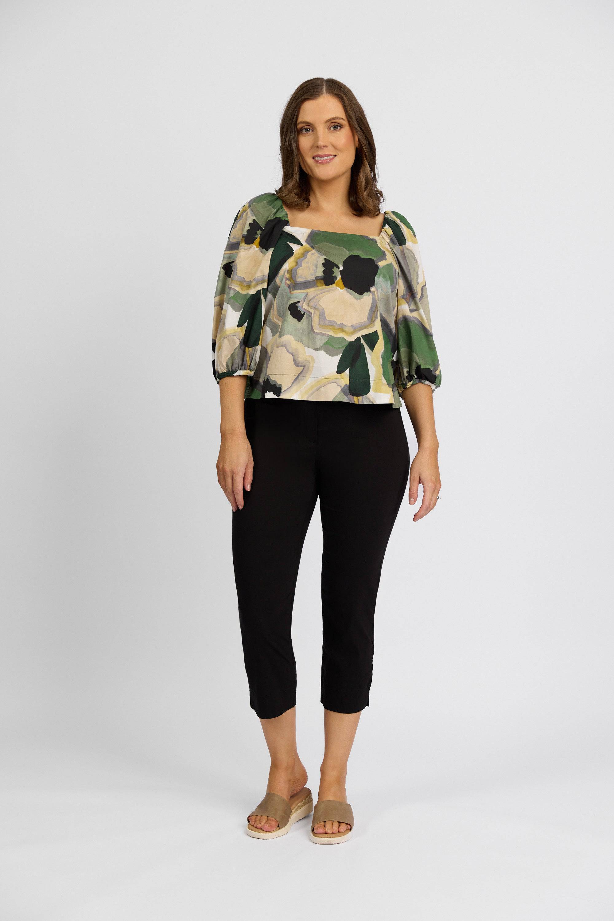 A woman with medium-length brown hair and a slight smile stands against a white background, wearing a floral puff-sleeve blouse, Vassalli Paris Crop Pant - Black crafted from stretch fabric, and beige slide sandals.