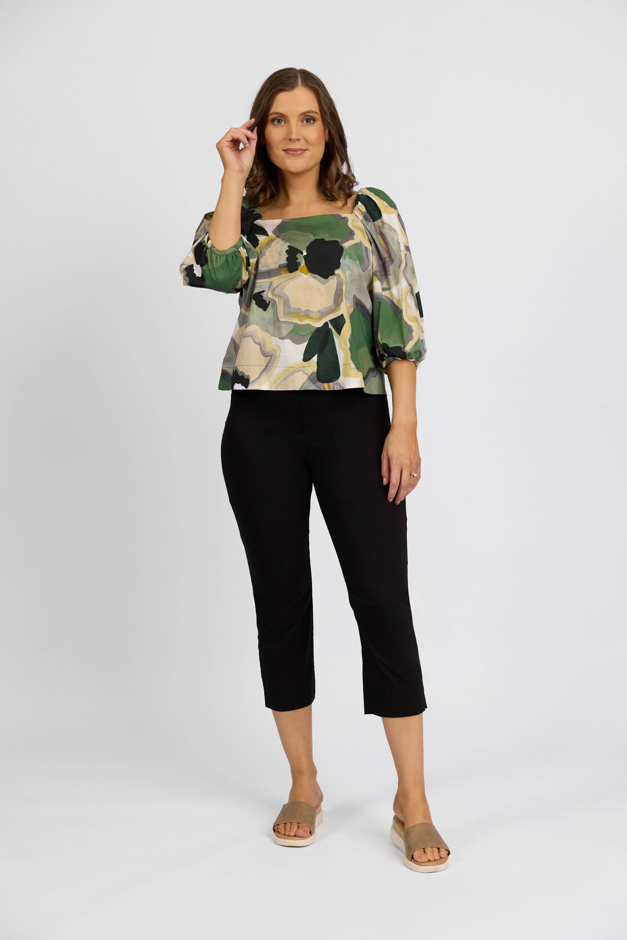 A woman stands against a white background, smiling in a floral green and beige top, Vassalli Paris Crop Pant - Black, and beige slide sandals. She holds one hand near her face. The outfit is made in New Zealand.
