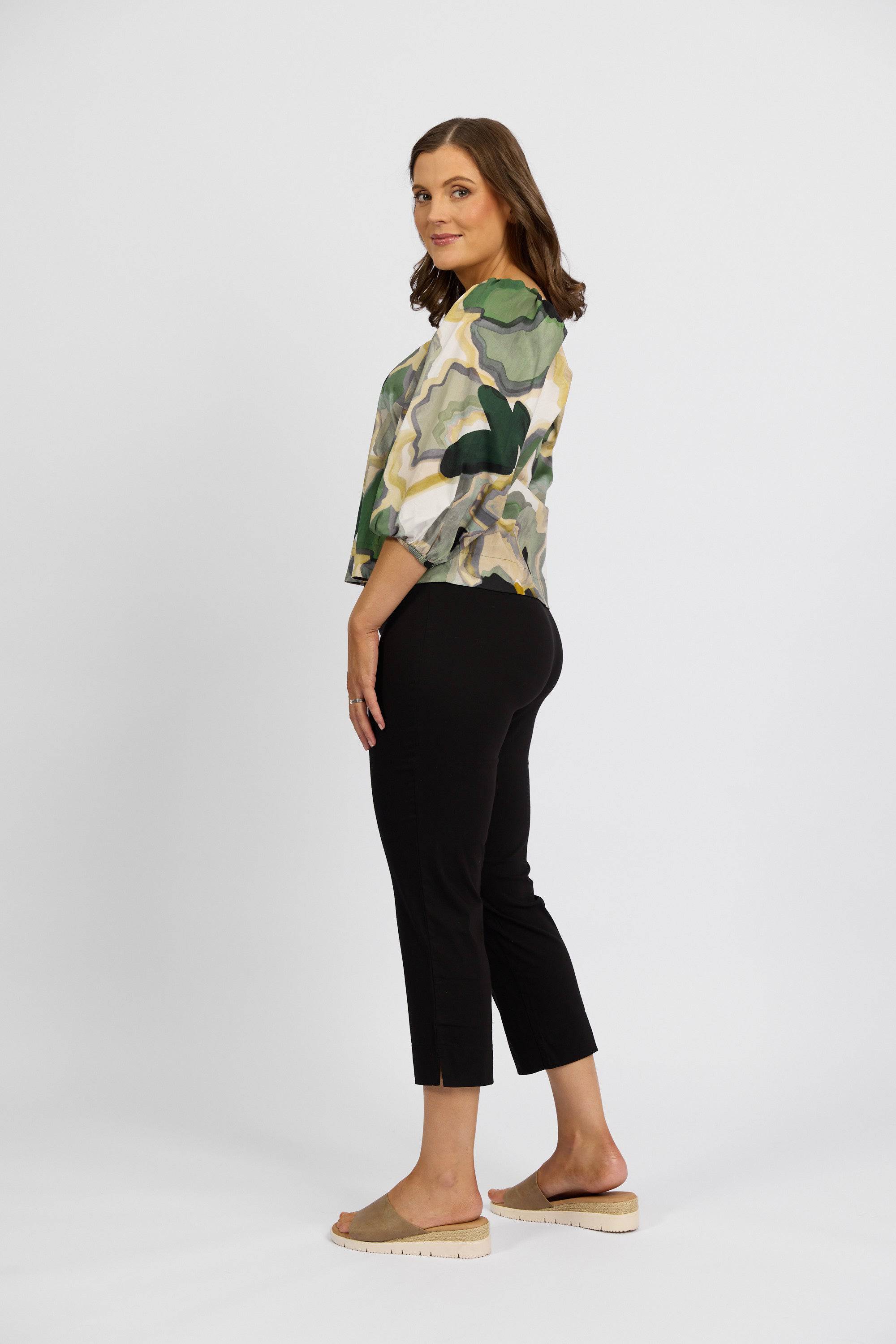 A brown-haired woman smiles over her shoulder, wearing a green and yellow patterned blouse, Vassalli Paris Crop Pant - Black, and beige slip-on shoes against a plain white background.