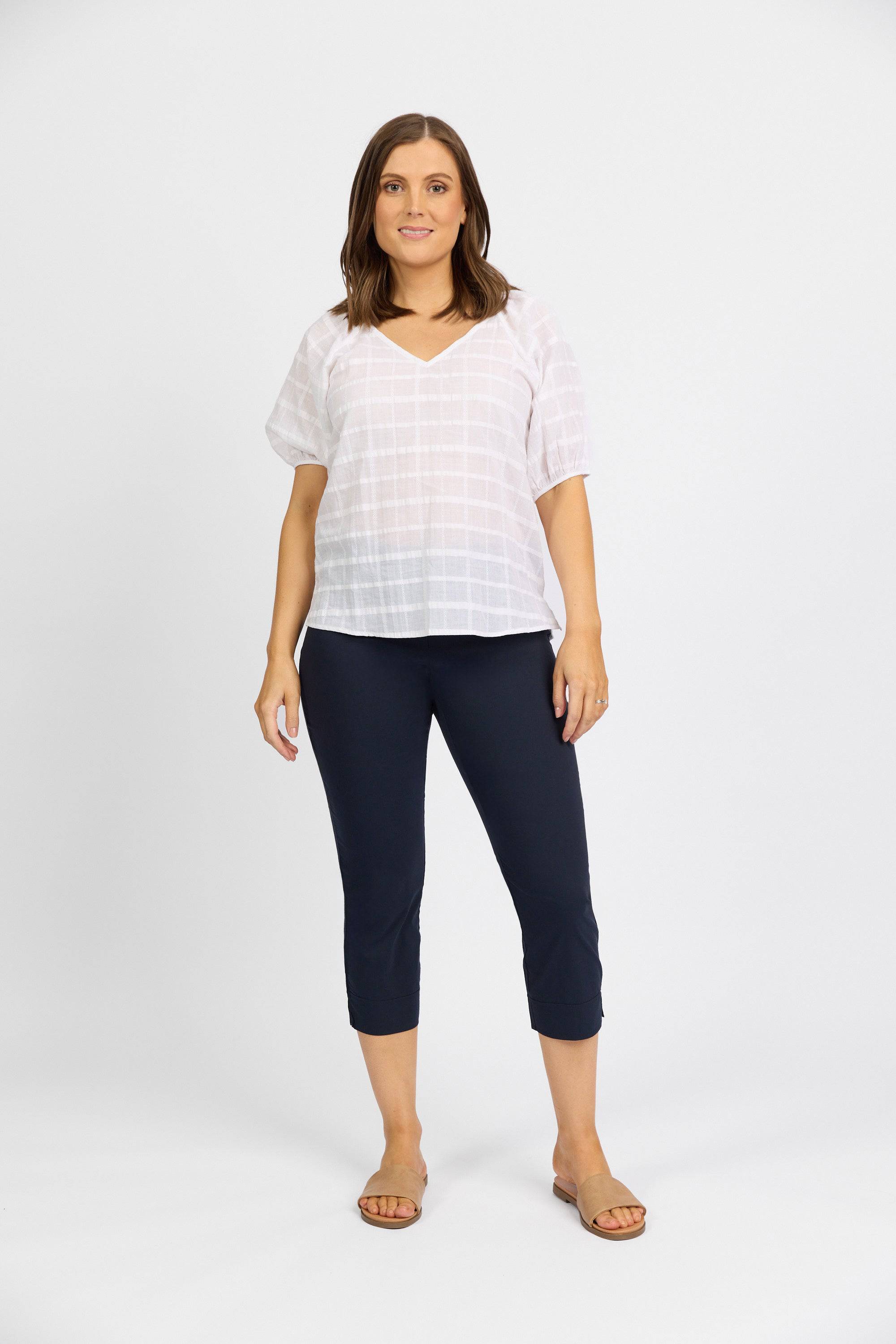 A woman stands against a white background wearing the Vassalli Paris Crop Pant - Inka in navy, paired with a loose white V-neck blouse and tan sandals. Her brown hair falls to her shoulders as she poses with a relaxed expression.