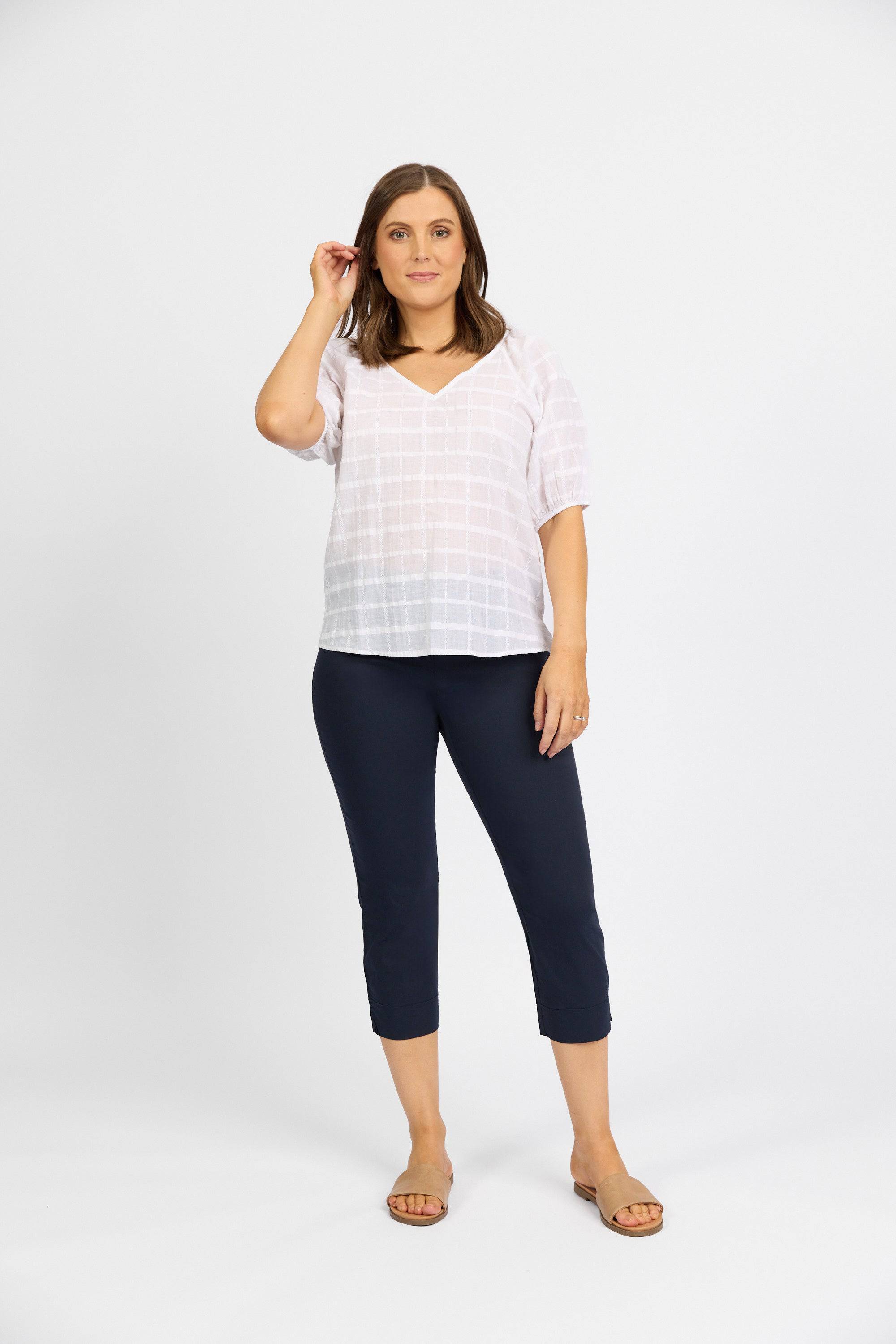 A woman with shoulder-length brown hair stands against a white background, wearing a white short-sleeve blouse, Vassalli Paris Crop Pant - Inka in navy from stretch fabric, and tan sandals. She touches her ear with one hand.