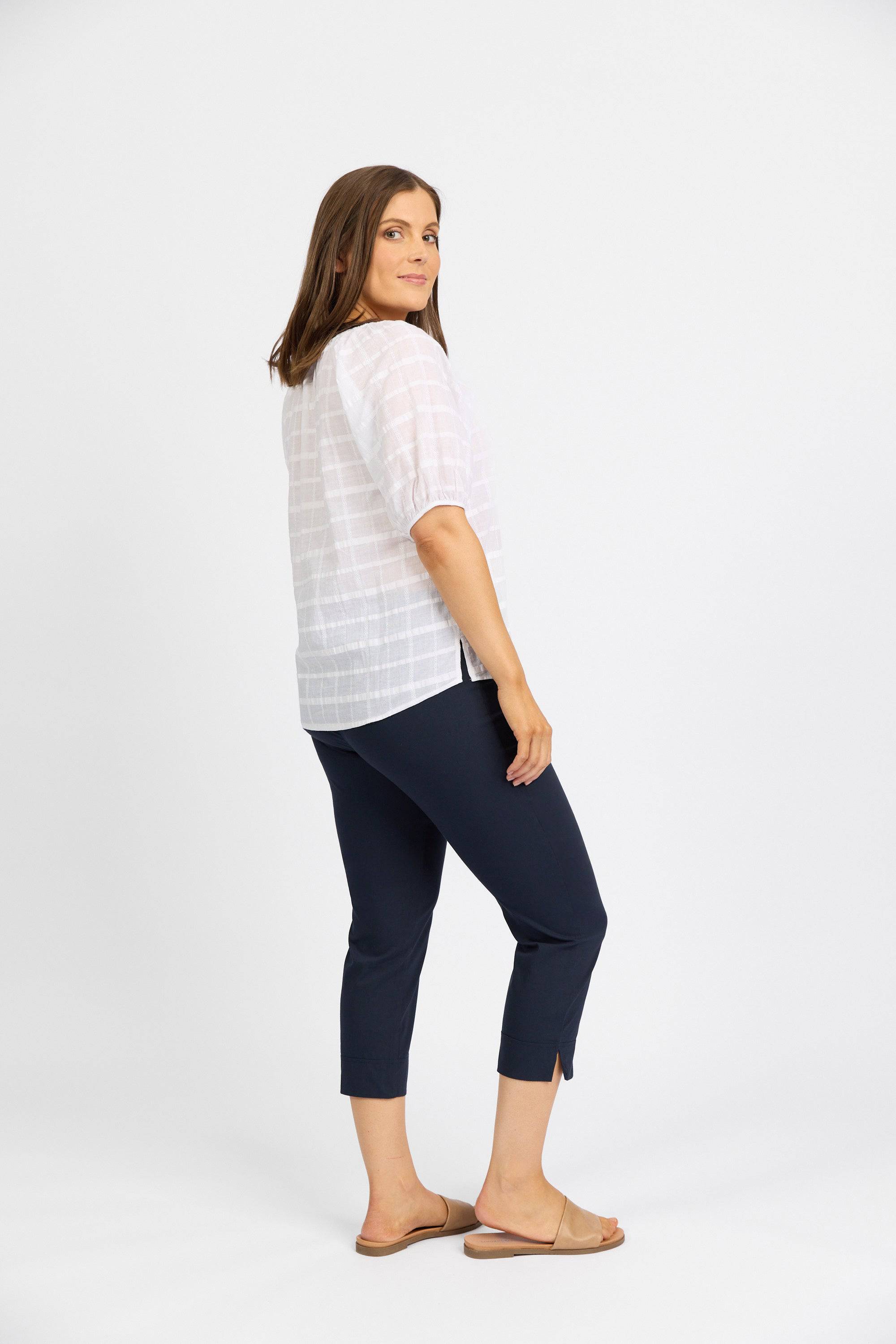 A woman stands sideways, looking over her shoulder. She wears a white blouse, tan slide sandals, and Vassalli Paris Crop Pant - Inka in dark navy stretch fabric with hem slits. The background is plain white.