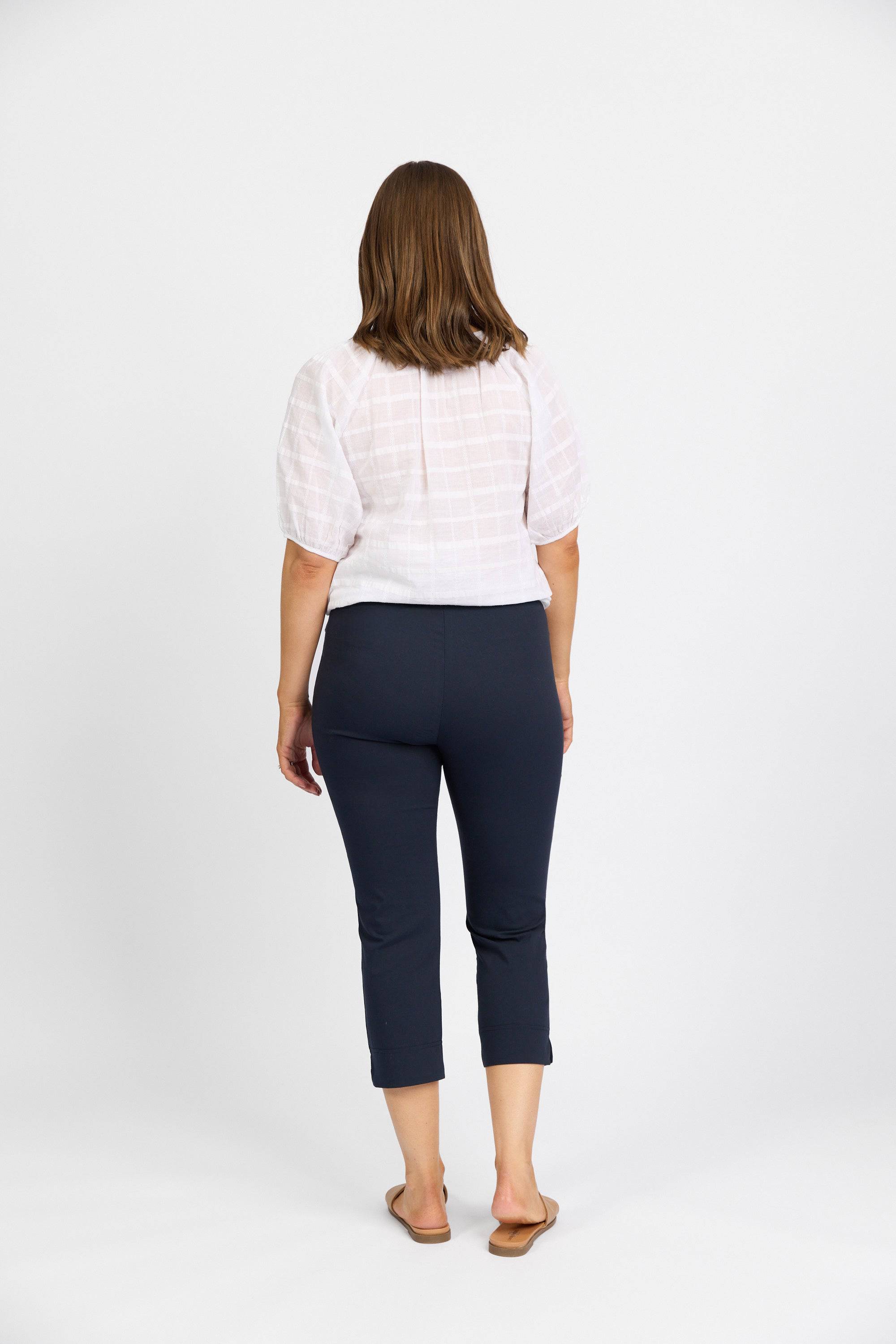 A woman with straight, shoulder-length brown hair stands with her back to the camera, wearing a white short-sleeve top and Vassalli Paris Crop Pant - Inka, paired with beige sandals against a plain white background.