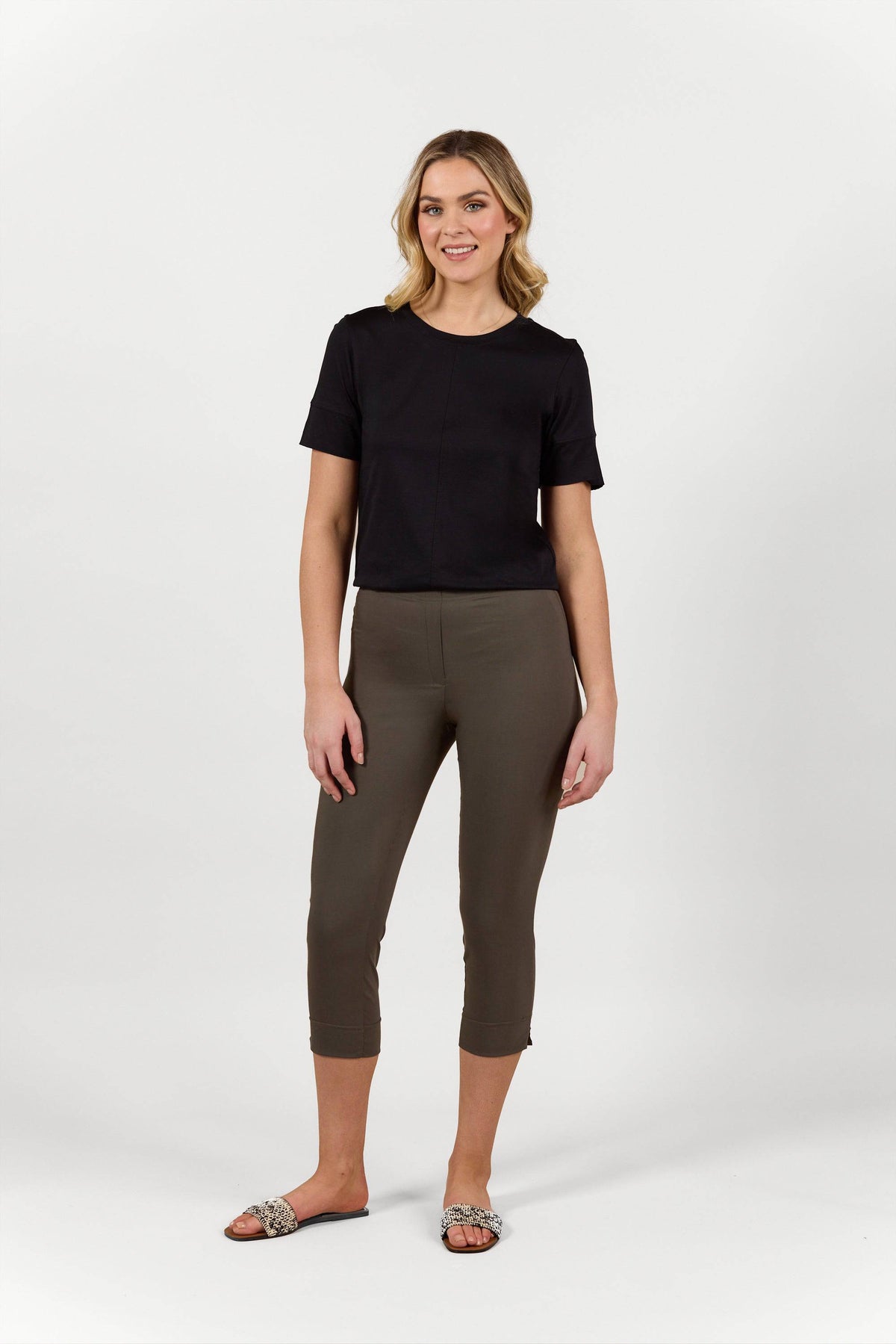 A smiling woman with blonde hair stands against a white background, wearing a black short-sleeve top, Vassalli's Paris Crop Pant in khaki, and slide sandals. One hand is relaxed by her side.