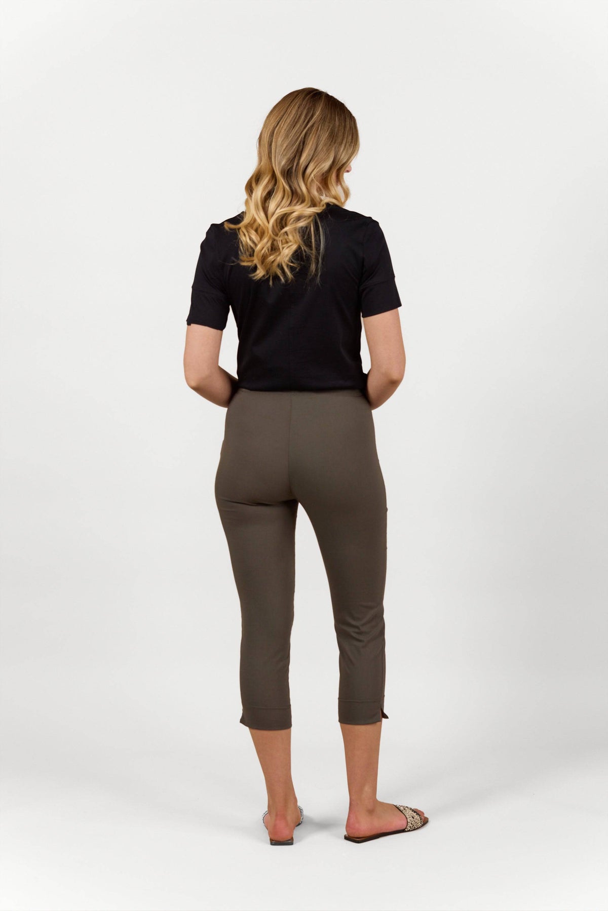 A woman with wavy blonde hair, seen from behind, wears a short-sleeved black top and Vassalli Paris Crop Pant in khaki. She stands against a plain white background and finishes the look with sandals.