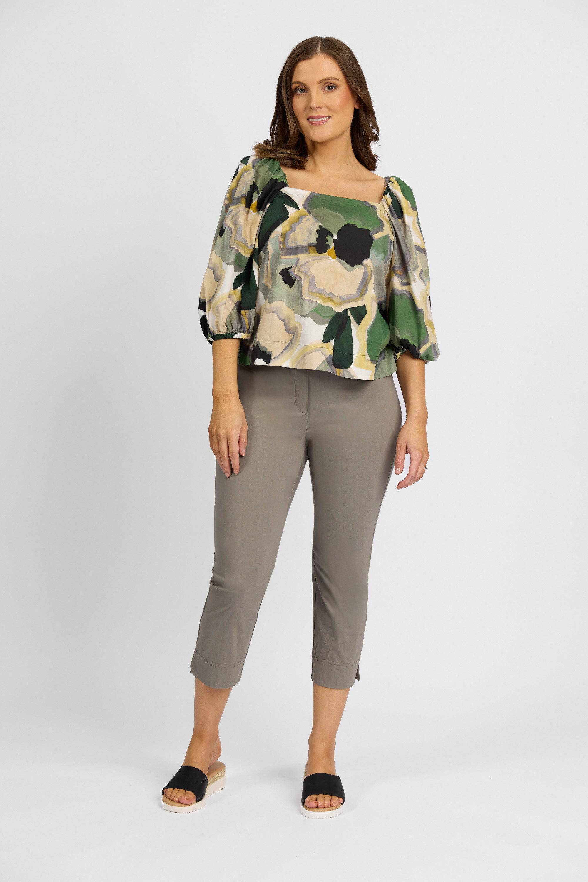A woman stands against a plain white background, smiling softly with one hand by her side. She wears a flowy floral blouse, Vassalli Paris Crop Pant in Latte, and black slide sandals.