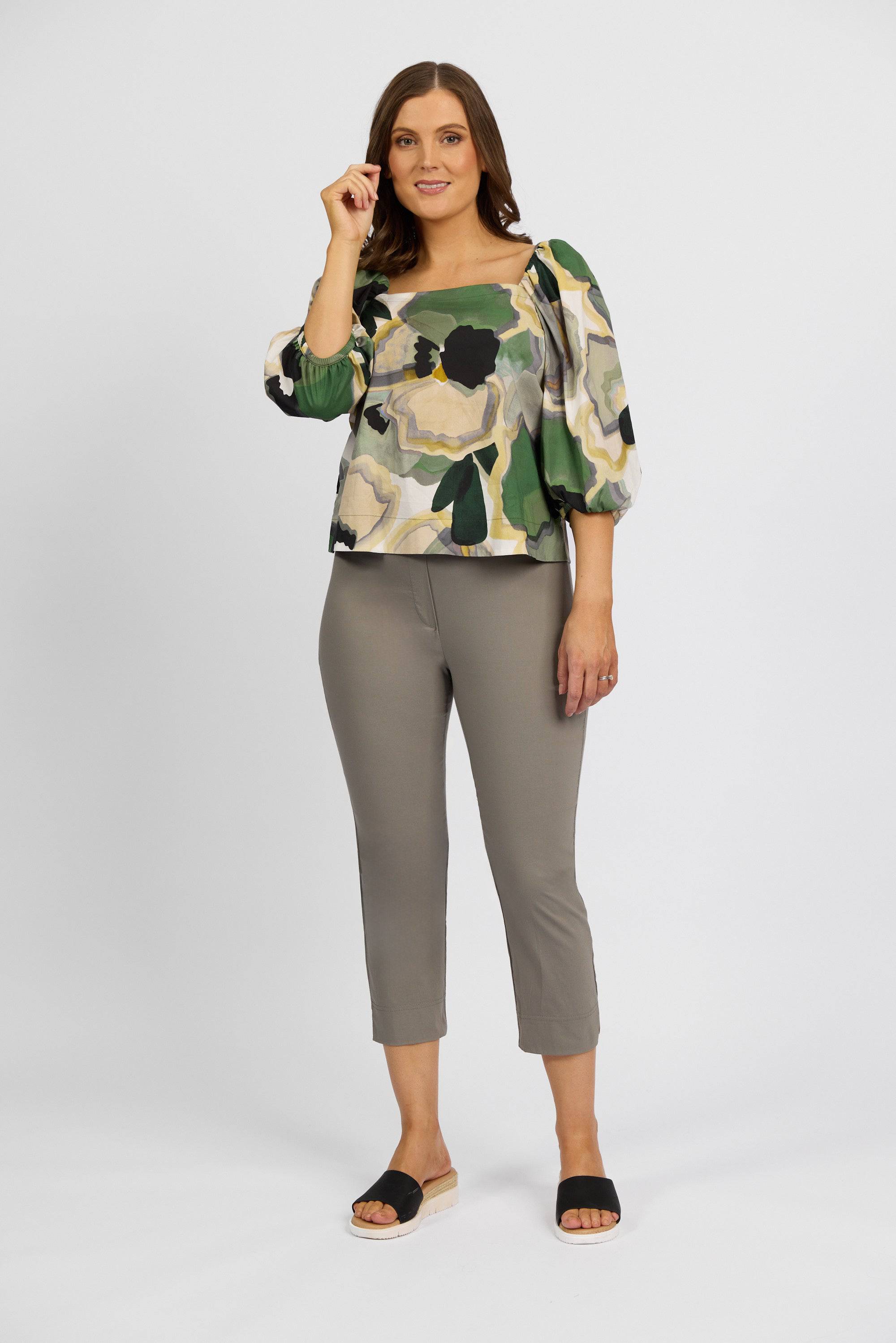 A woman stands against a plain background, smiling and touching her hair, wearing a green and beige floral puffed-sleeve blouse, Vassalli Paris Crop Pant - Latte, and black slide sandals.