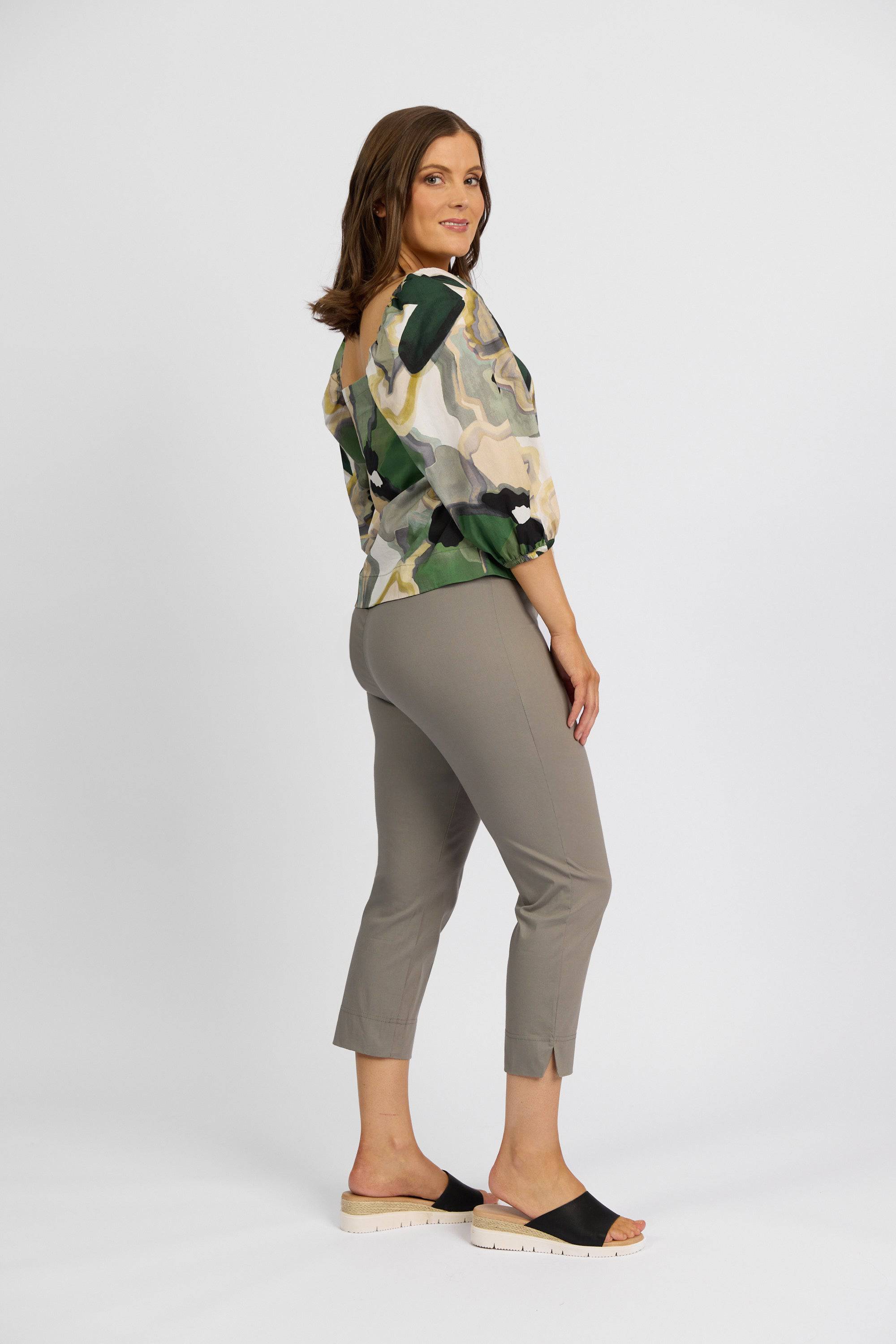 A woman stands sideways, smiling, wearing a patterned off-shoulder blouse with green and beige tones, Vassalli Paris Crop Pant - Latte, and black open-toe slide sandals against a plain white background.