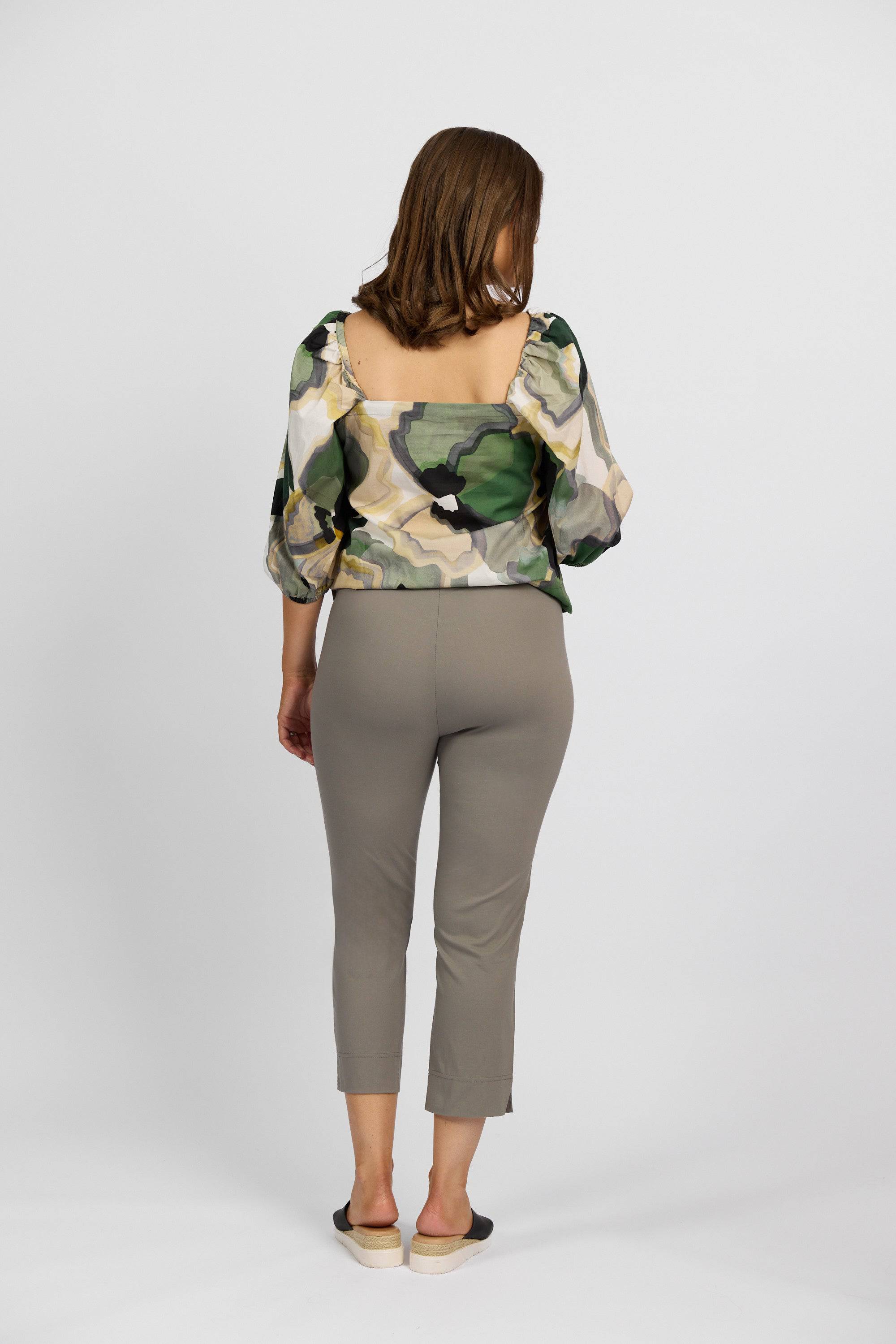 A woman with shoulder-length hair faces away, wearing a sheer green, cream, and beige patterned blouse from the Paris Collection, Vassalli Paris Crop Pant in Latte, and black slip-on shoes against a plain white background.