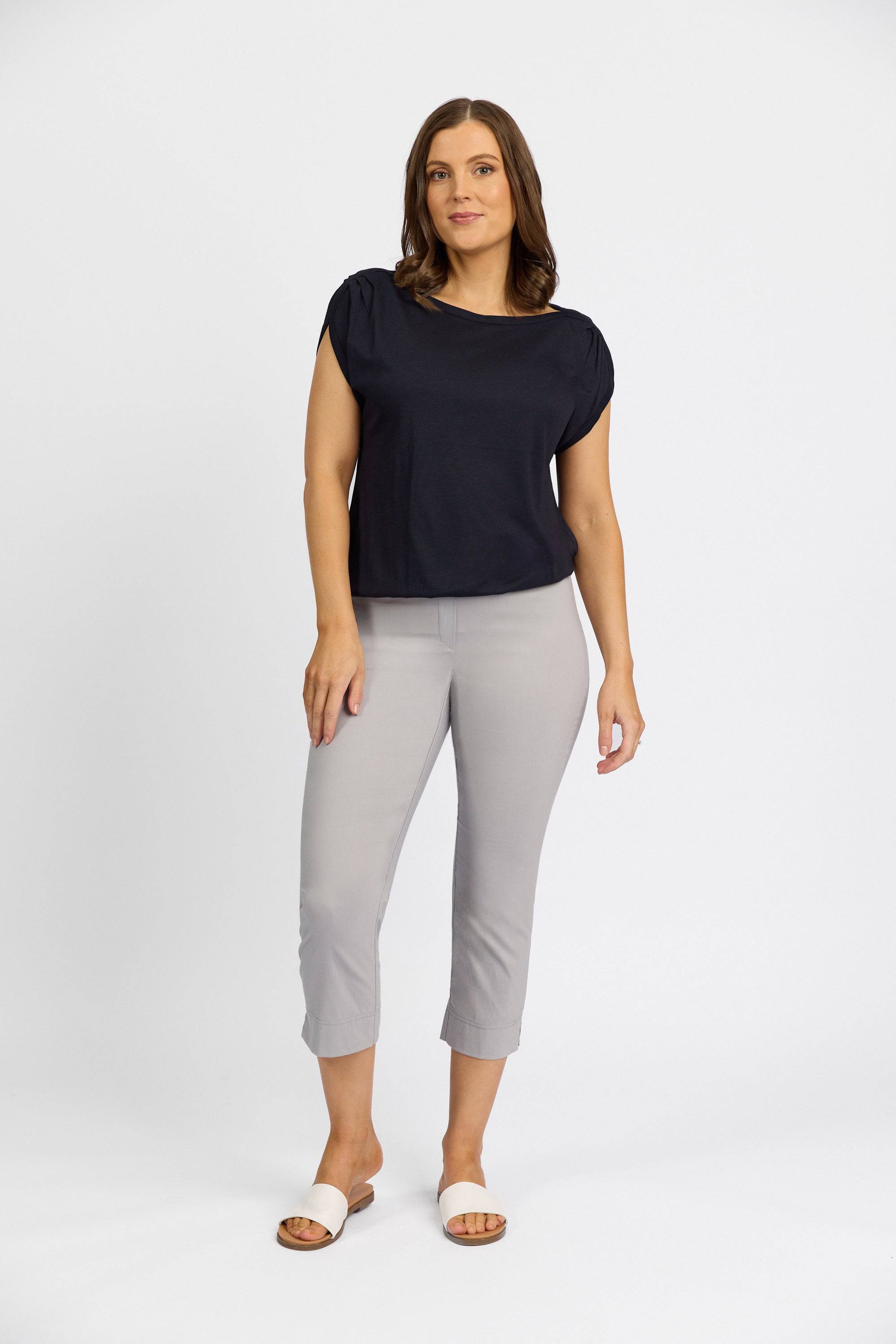 A woman with shoulder-length brown hair poses confidently against a white background, wearing a black short-sleeve top, Vassalli Paris Crop Pant - Silver, and white slide sandals.