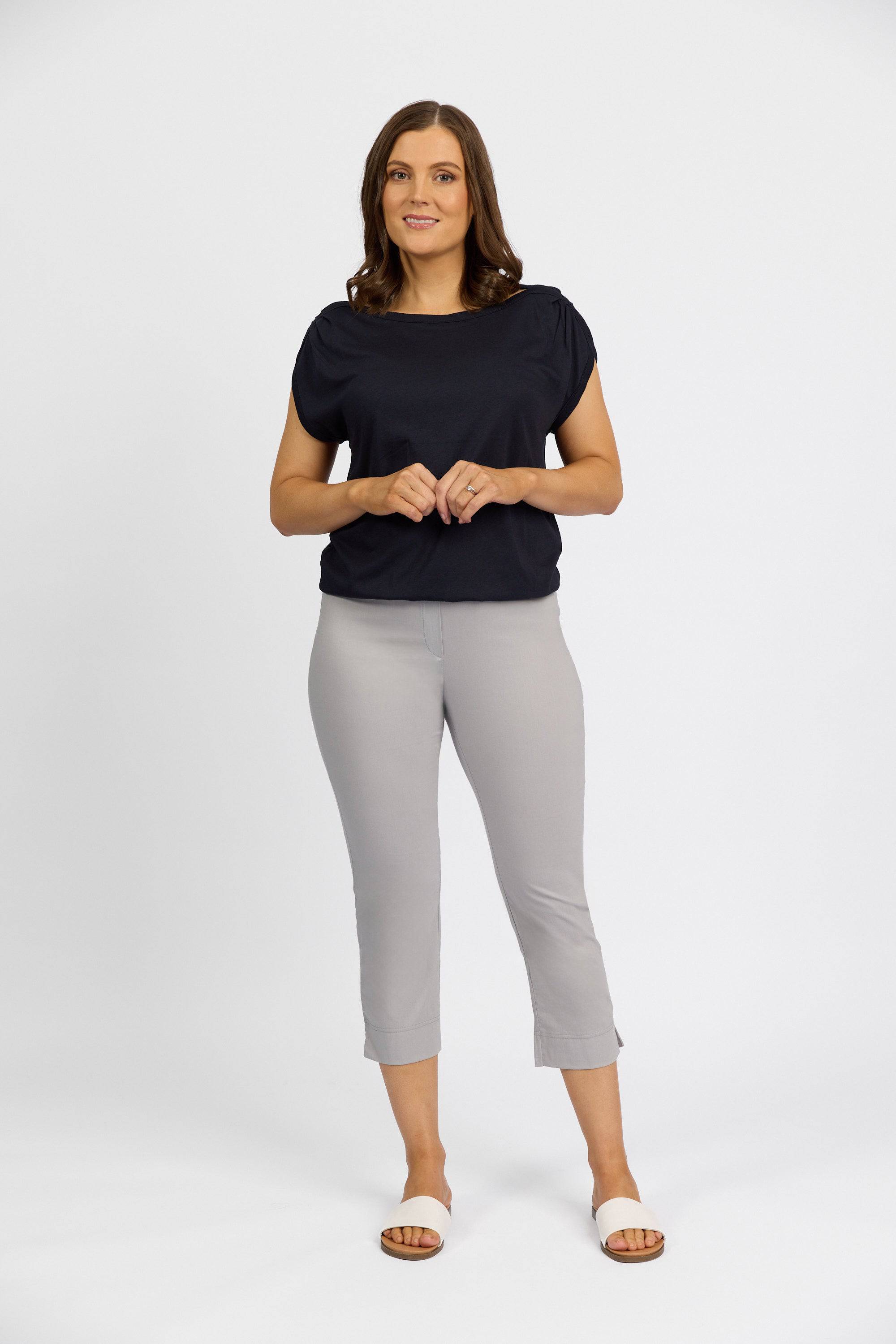 A woman with brown hair stands facing forward, smiling softly with hands clasped. She wears a black short-sleeve top and Vassalli Paris Crop Pant in Silver, paired with white slide sandals, against a plain white background.