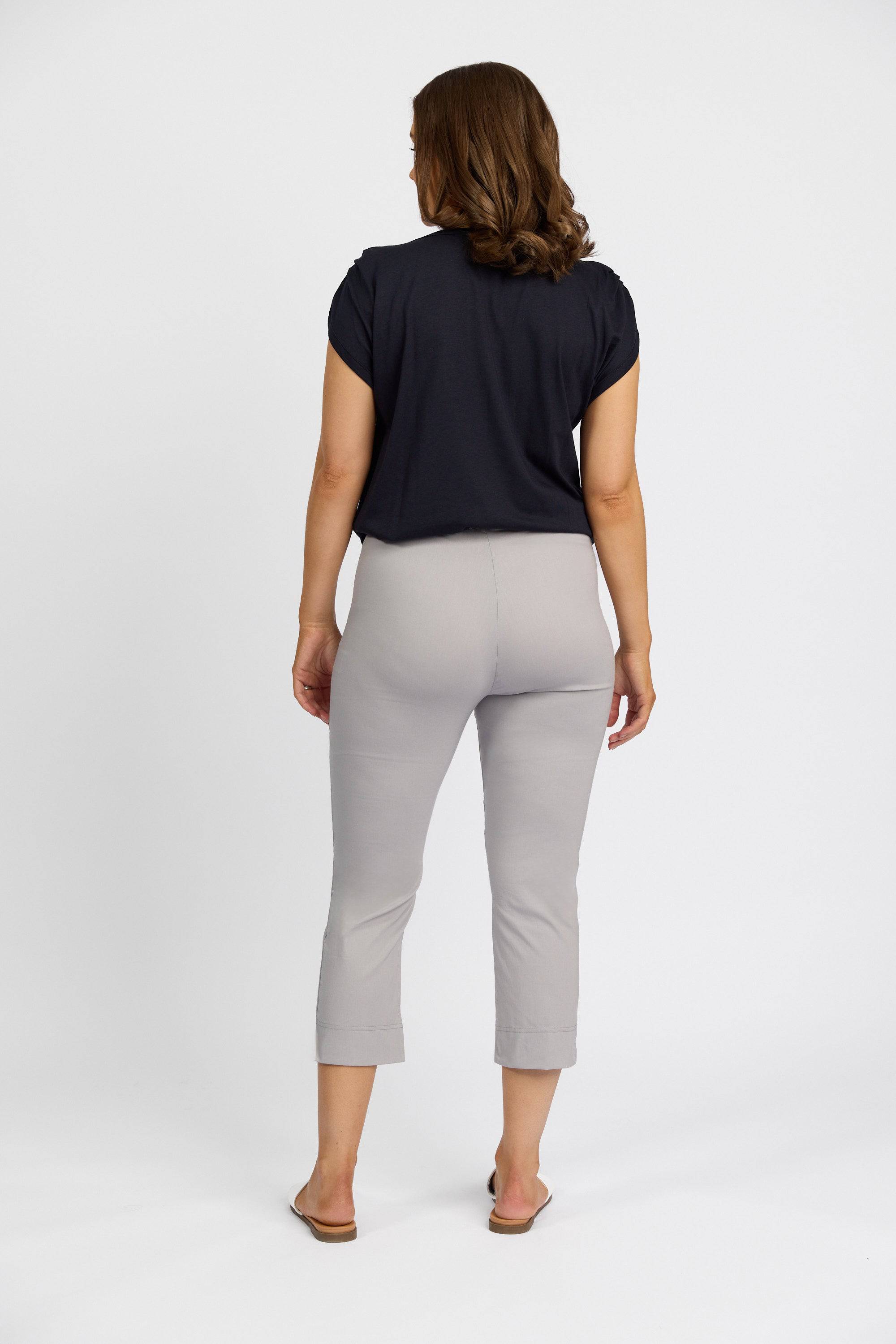 A woman with wavy brown hair, wearing a black short-sleeve top and Vassalli Paris Crop Pant - Silver made from stretch fabric in New Zealand, stands facing away against a plain white background.