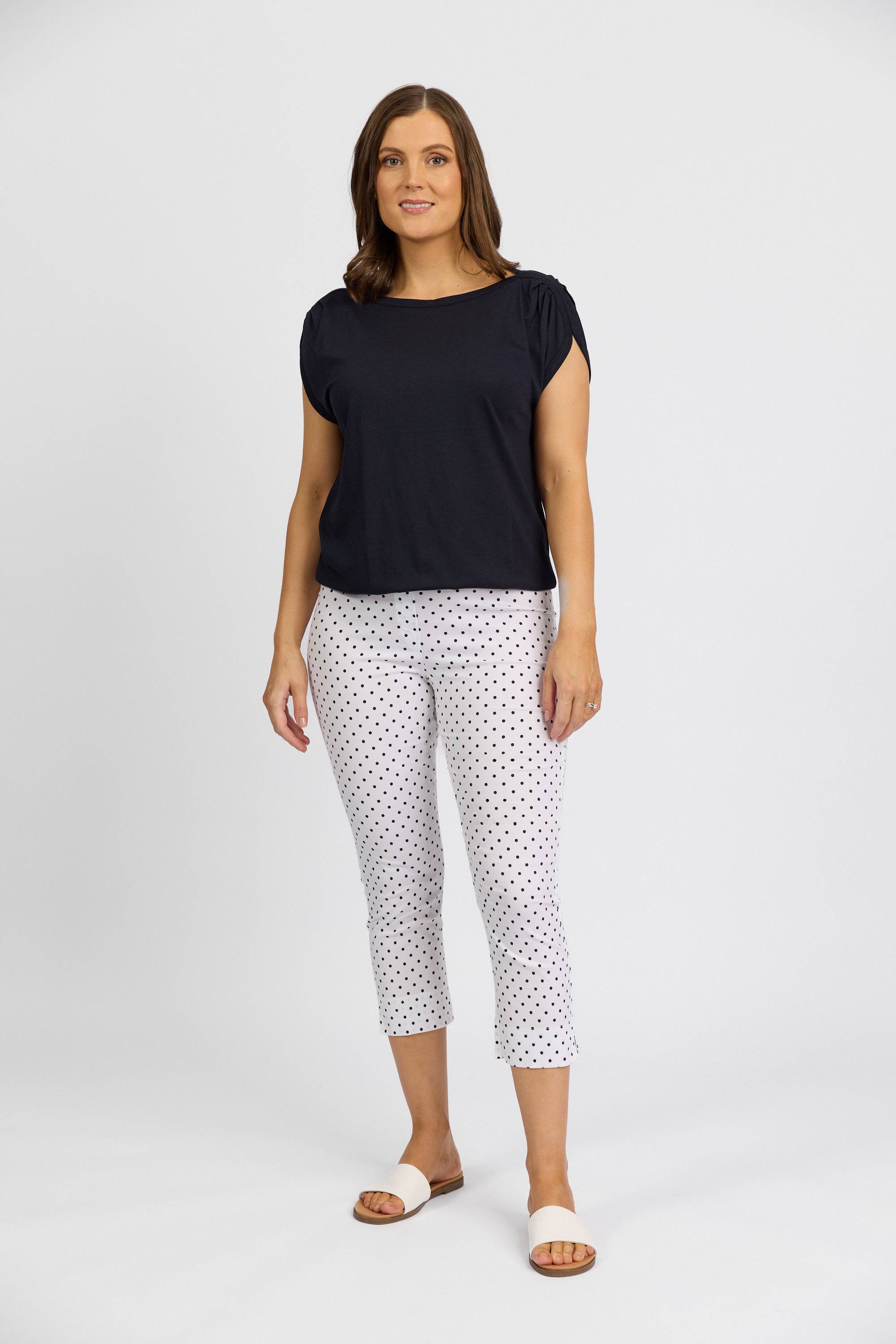 A woman with medium-length brown hair and a relaxed expression wears a black short-sleeve top, Vassalli Paris Printed Crop Pant - Spot, and white slide sandals against a plain white background.