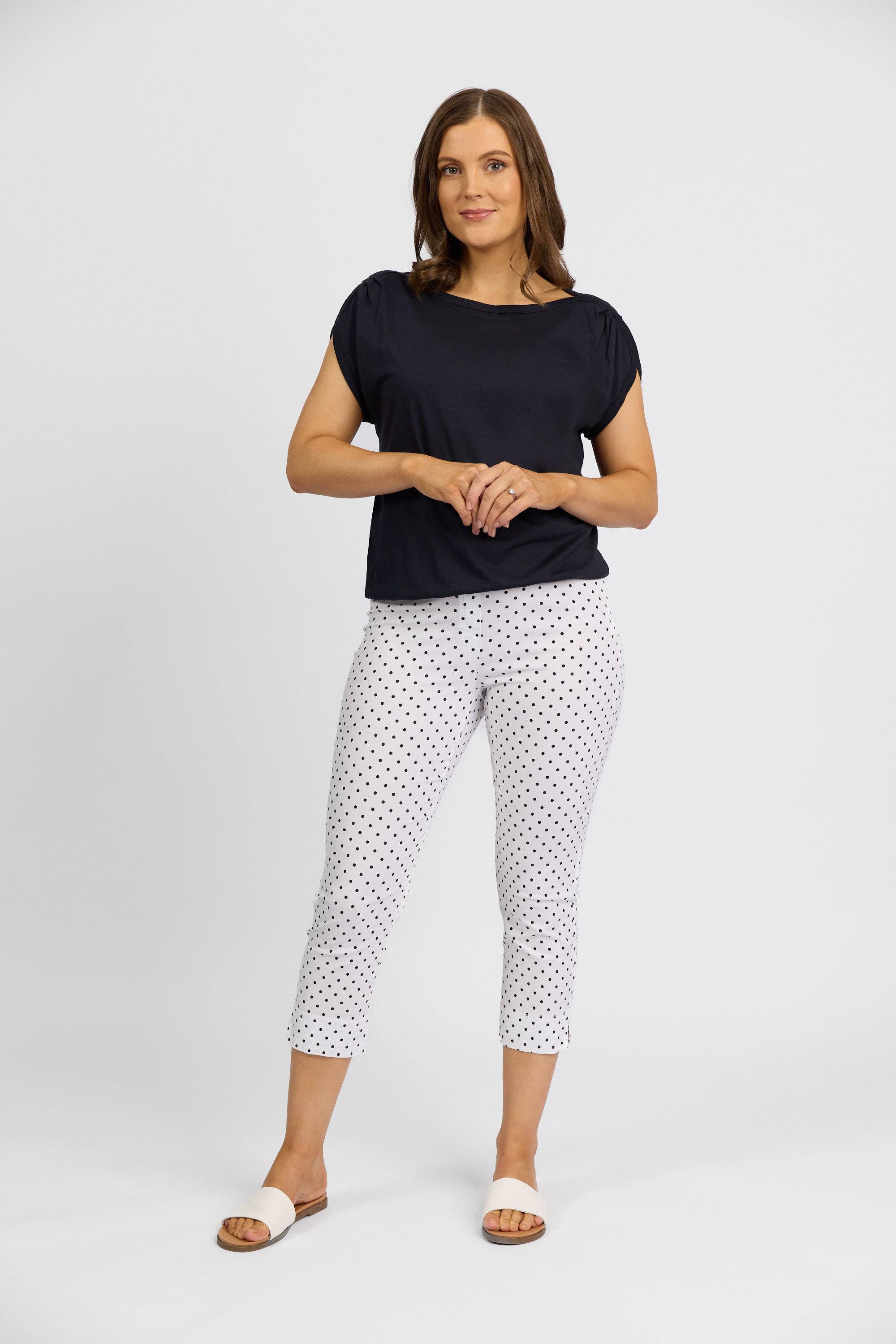 A woman with medium-length brown hair and a confident posture wears a black short-sleeve top, Vassalli Paris Printed Crop Pant - Spot in stretch fabric, and beige slide sandals against a plain background.