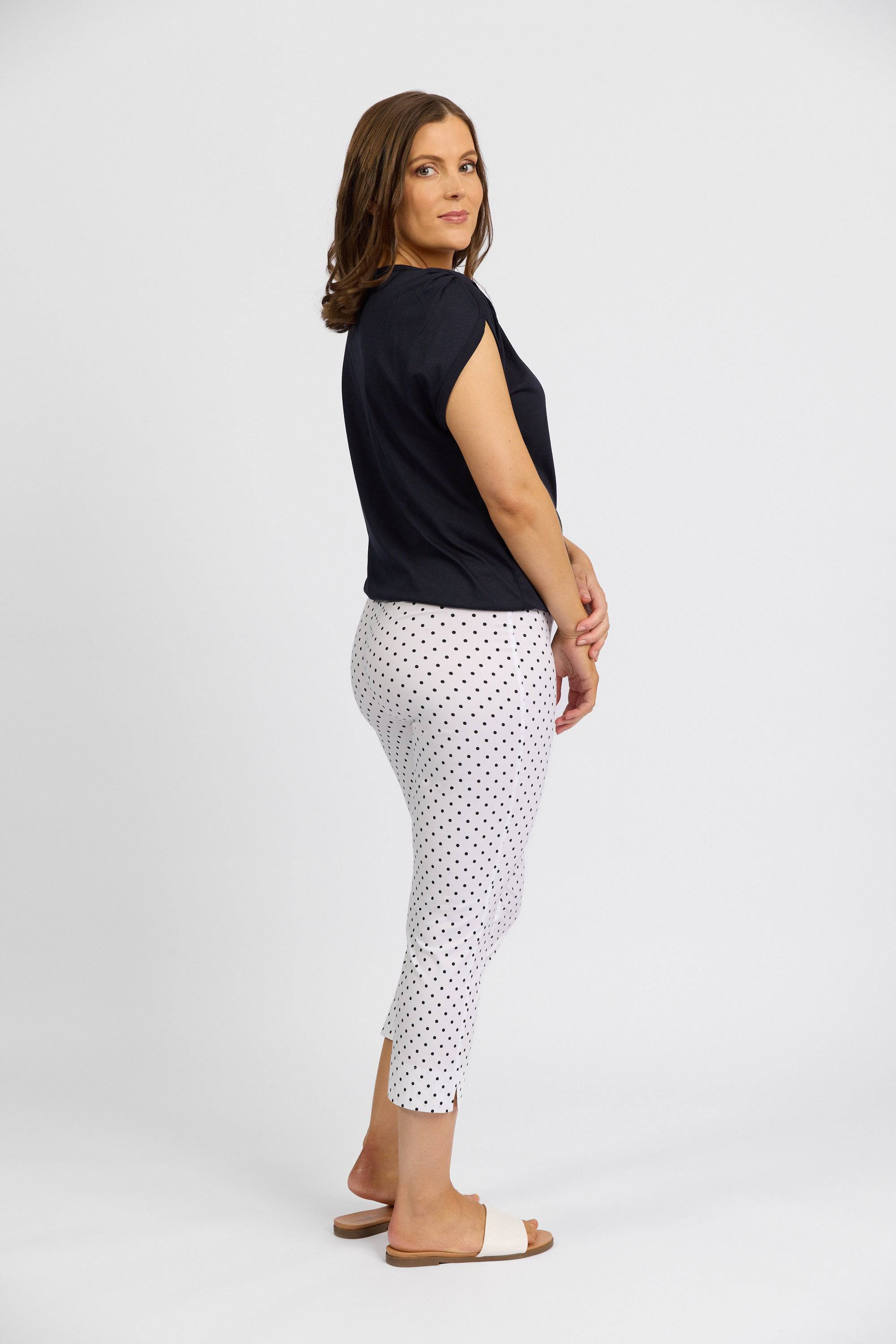 A woman stands sideways, looking at the camera, wearing a black t-shirt and Vassalli’s Paris Printed Crop Pant - Spot—white with black polka dots—paired with white sandals, against a plain white background.