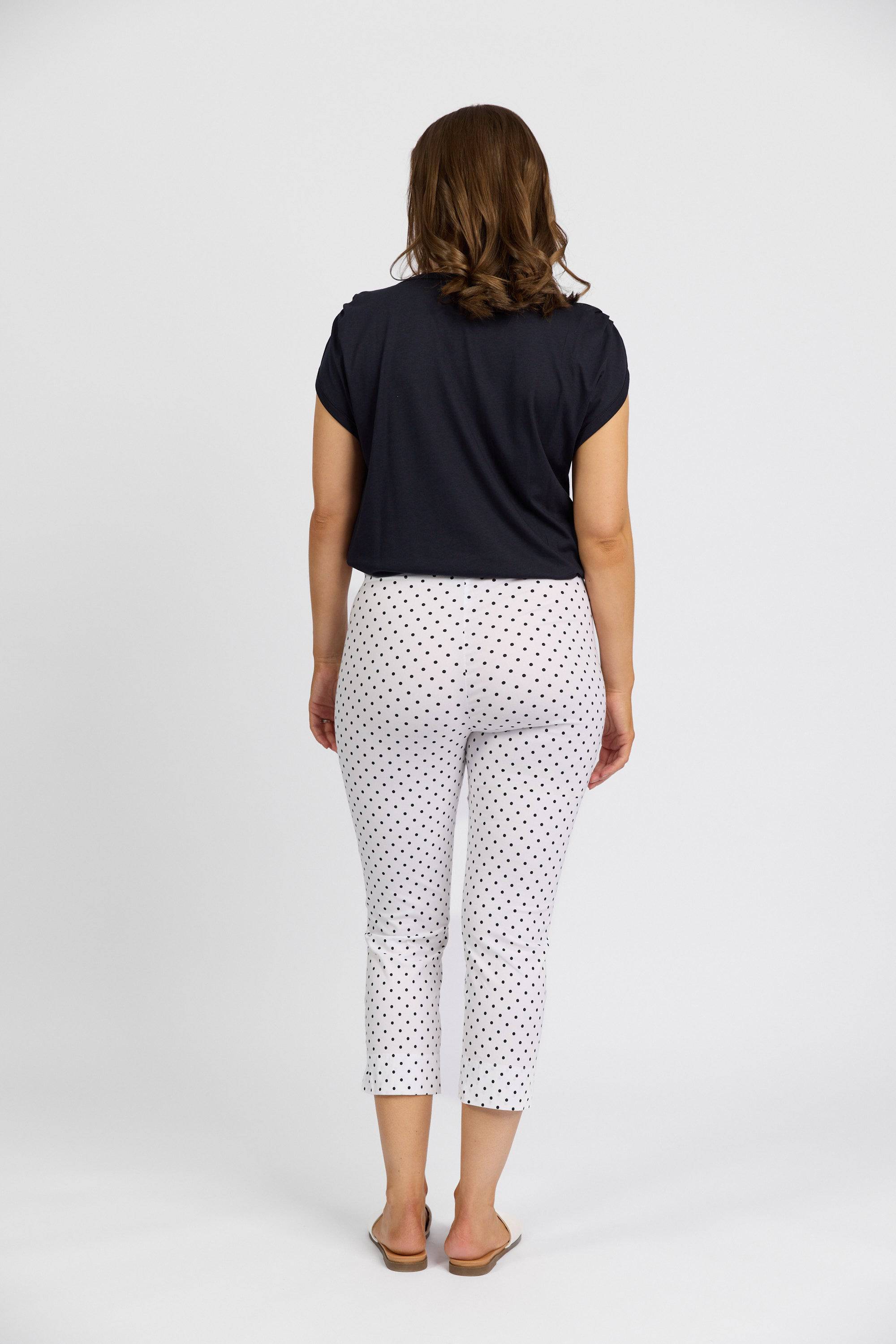 A woman faces away from the camera in a short-sleeve black top and Vassalli Paris Printed Crop Pant - Spot, featuring white fabric with small black dots. She finishes the look with flat sandals against a plain white background.
