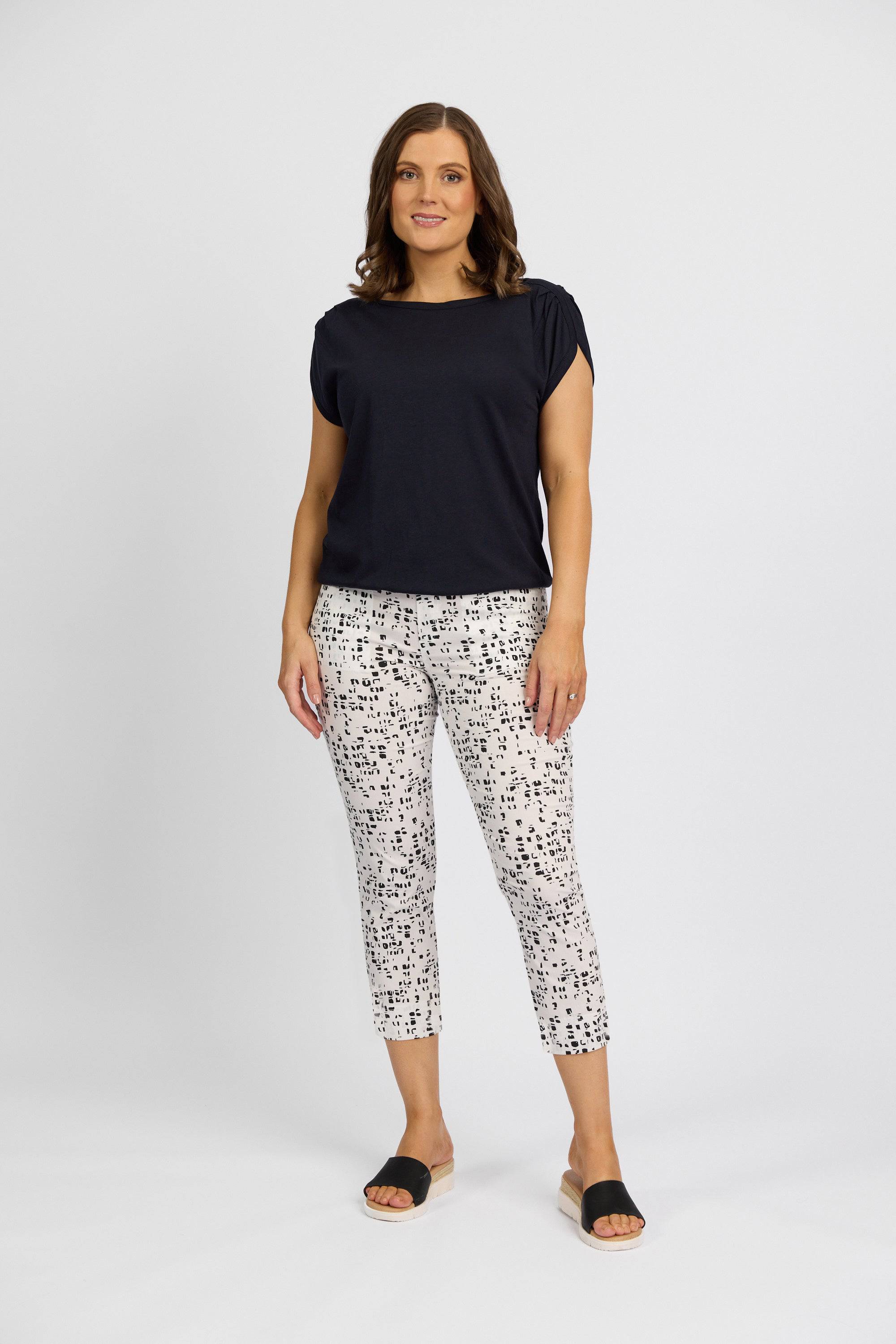 A woman with shoulder-length brown hair smiles slightly while wearing a black short-sleeve top, Vassalli Paris Printed Crop - Tonic white ankle-length pants with a black abstract pattern, and black slide sandals against a plain white background.