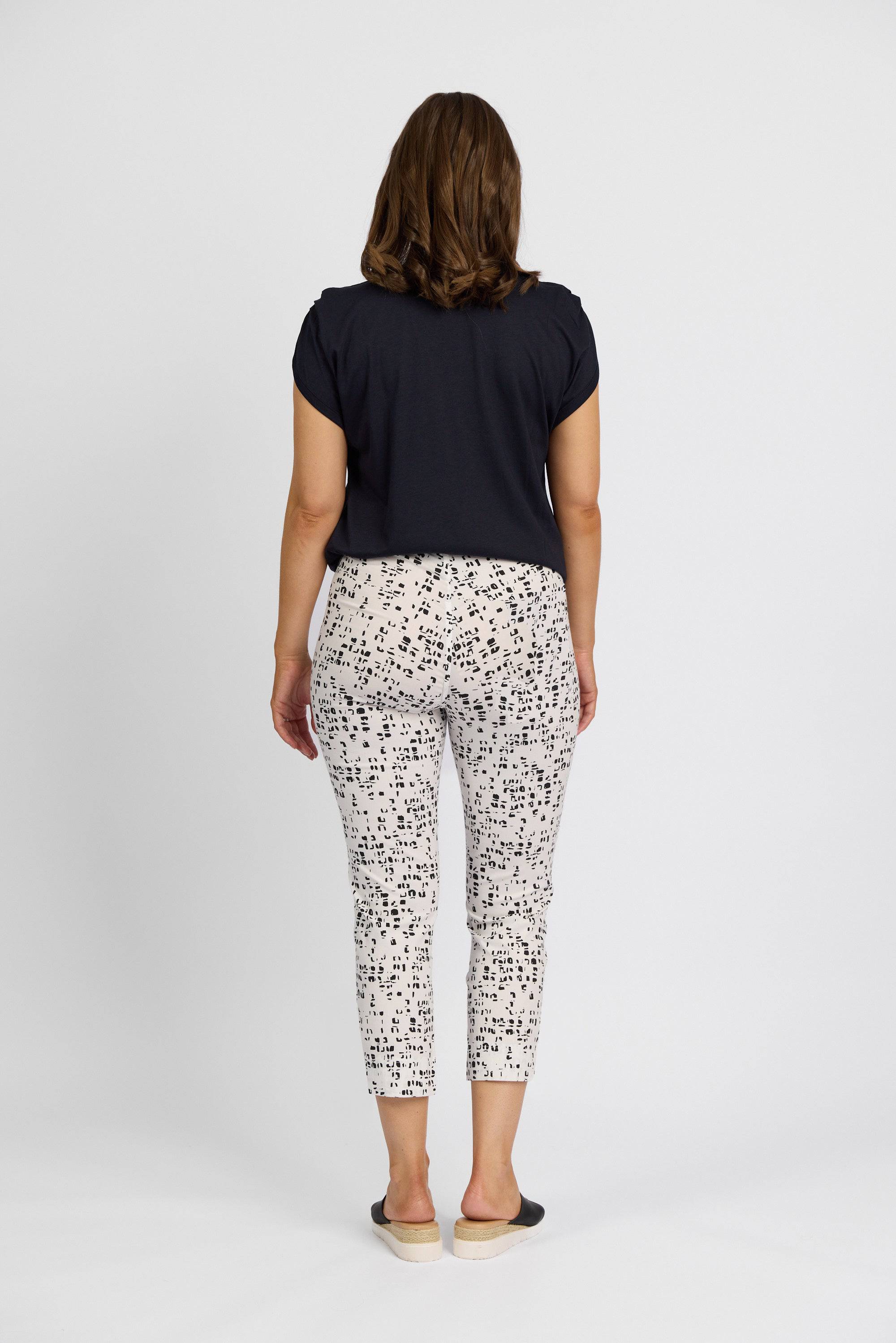A woman faces away from the camera wearing a black short-sleeve top and Vassalli Paris Printed Crop - Tonic pants with a black abstract print, paired with black slip-on shoes, against a plain white background.